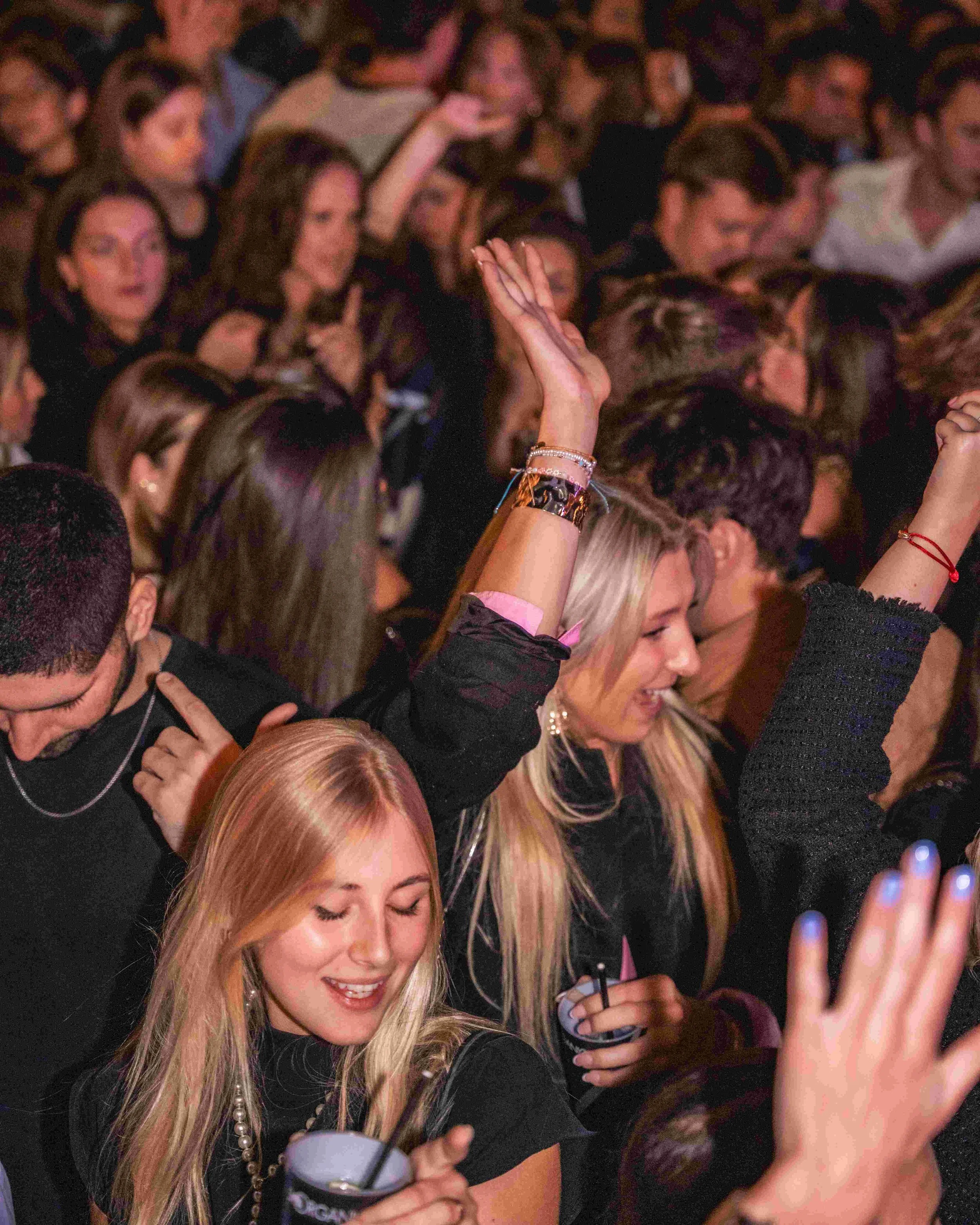 A crowded party scene with numerous people closely gathered, with some people raising their hands and holding drinks. A woman with long blonde hair is smiling in the foreground.