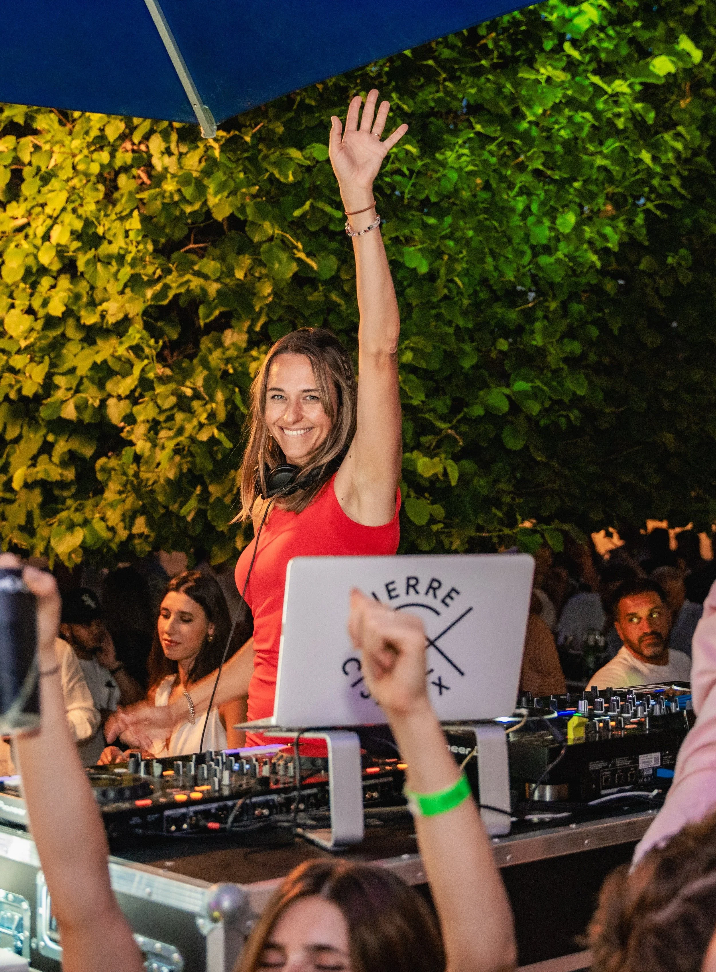 A young woman DJ in a red top with headphones around her neck, smiling and raising her right hand, at a lively outdoor event with a crowd and DJ equipment, illuminated by green and yellow lights, with a leafy background.