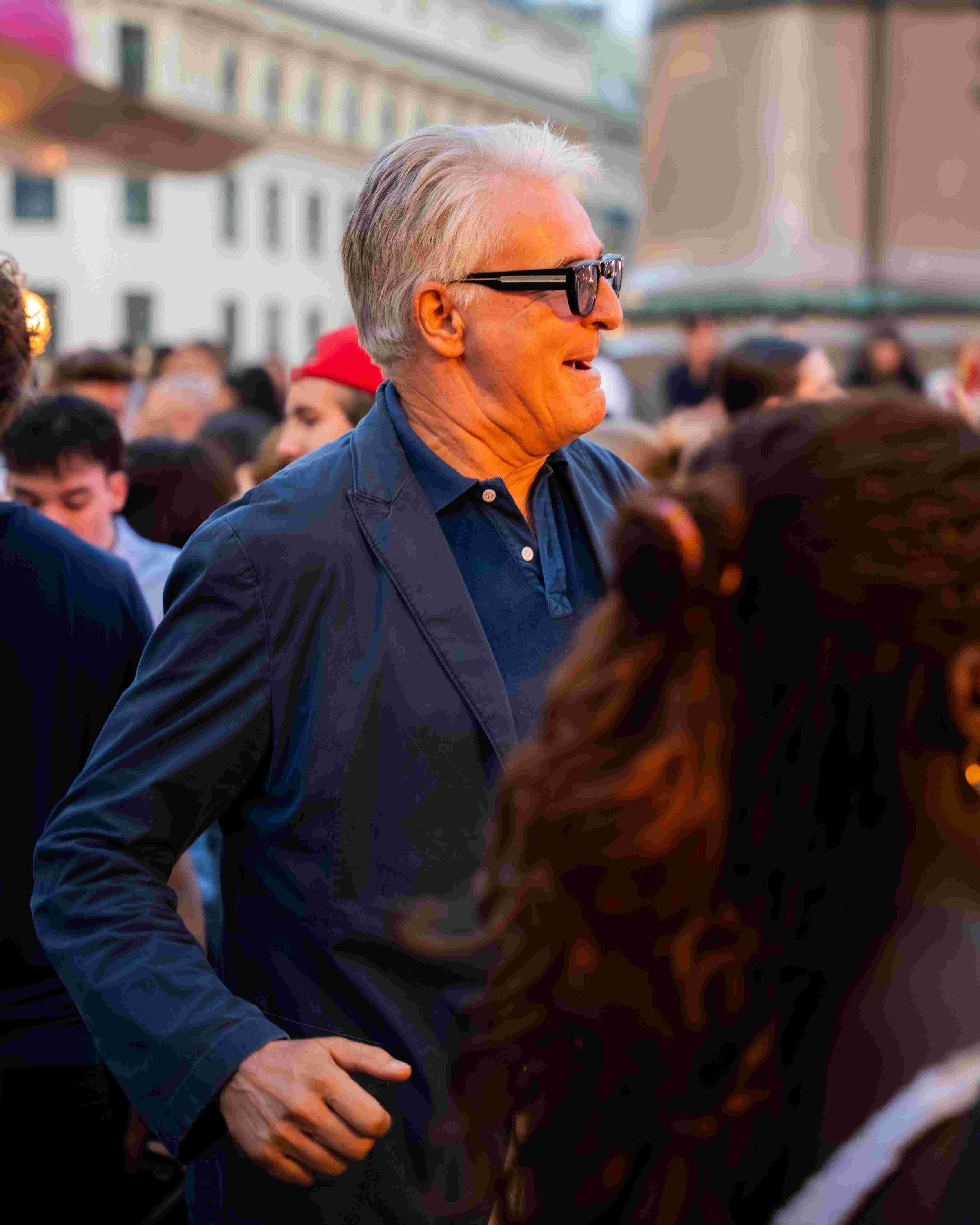 A middle-aged man with gray hair, wearing black glasses and a blue jacket, smiling and interacting with others during an outdoor gathering or event in a city environment.