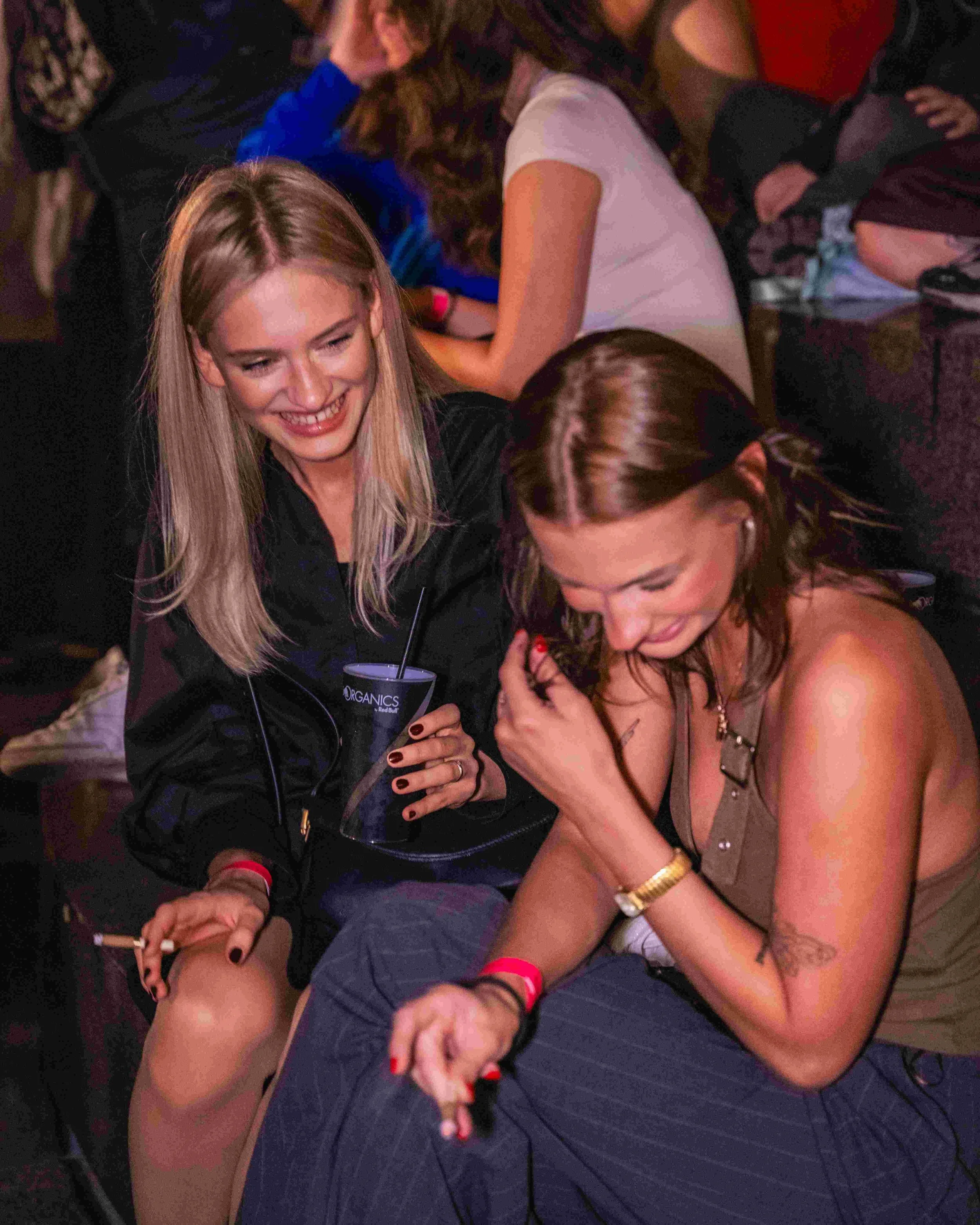 Two women are hugging and smiling at a party or social gathering, sitting close together on a dark-colored floor, with other people around them.