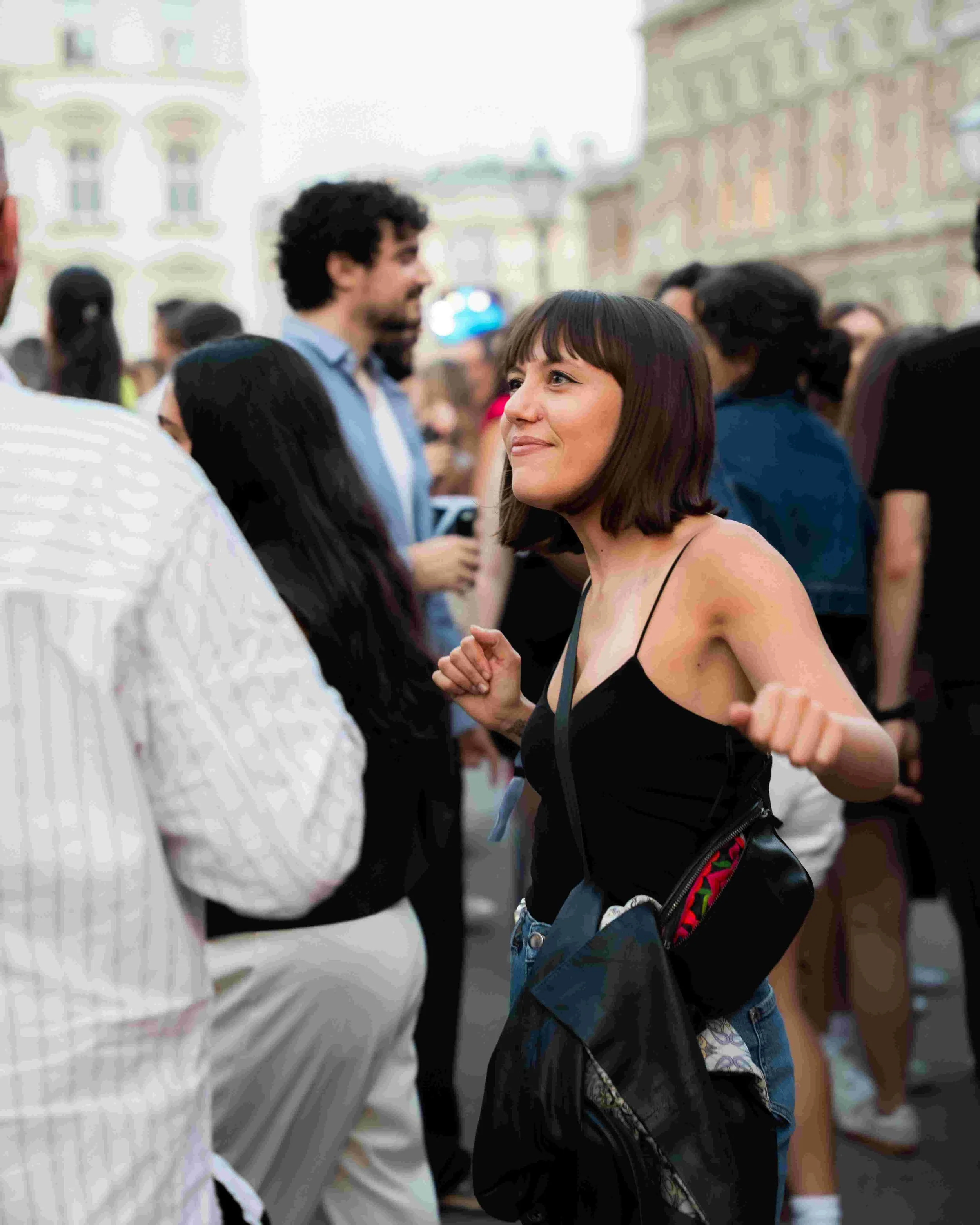 A woman with short dark hair wearing a black spaghetti strap top and denim shorts smiling and dancing at an outdoor event with a crowd of people and historic buildings in the background.