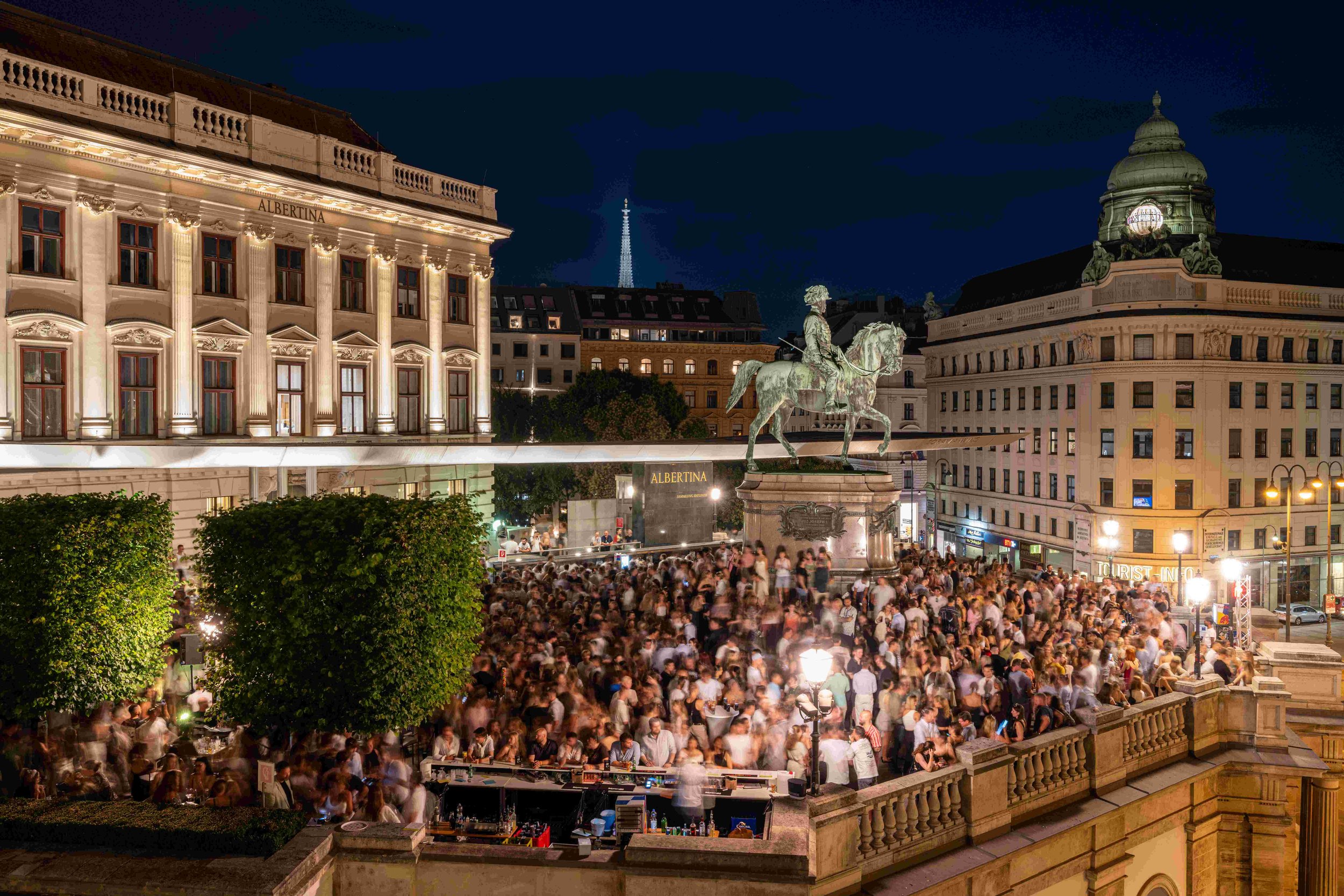 Night scene of Alexanderplatz in Berlin, Germany, with a large crowd gathered in the square. Prominent buildings are illuminated, including the Albertina building on the left, a statue of a person on a horse in the center, and the Fernsehturm (TV tow