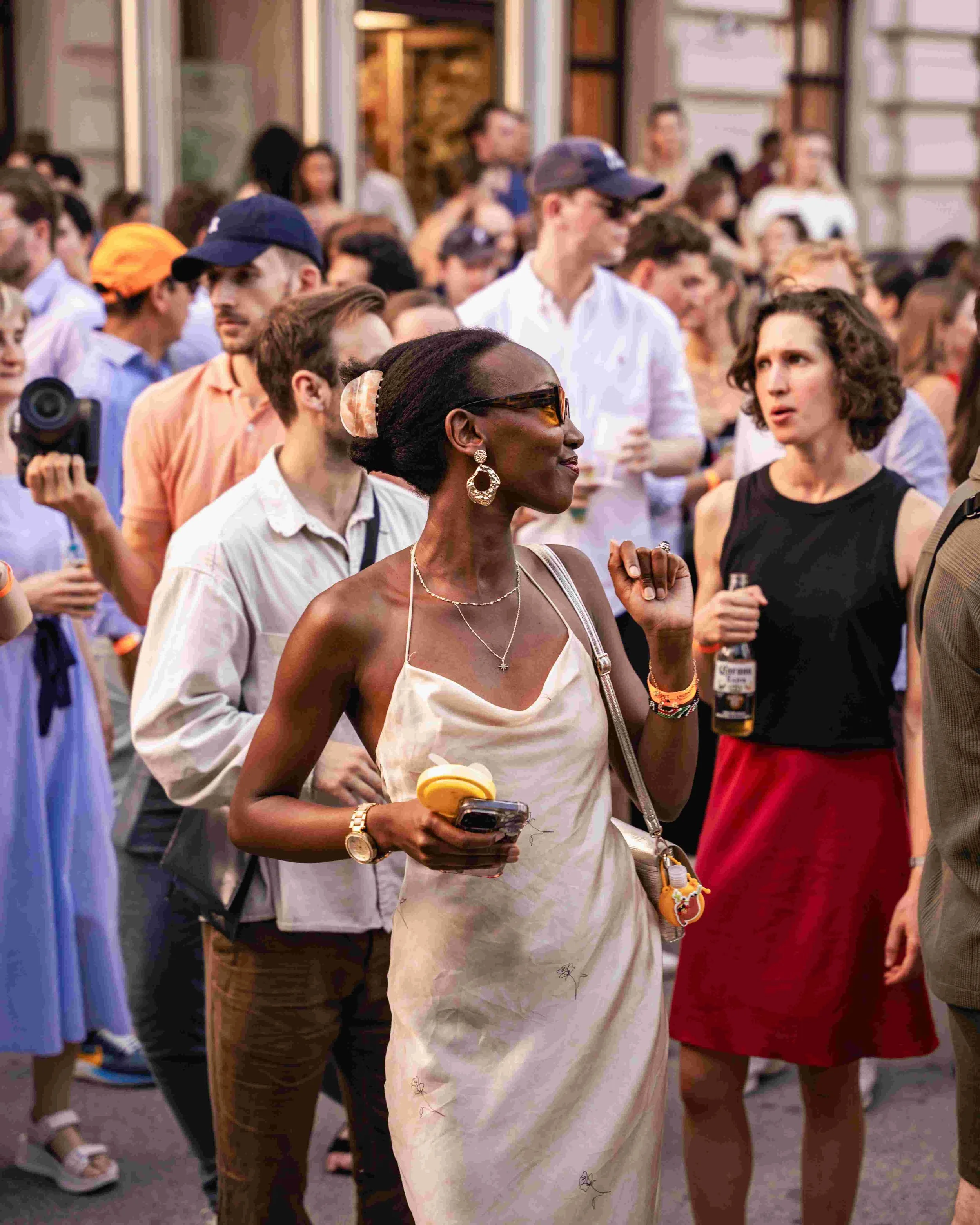 A diverse group of people, including a woman with earrings and sunglasses and a woman with curly hair, are gathered at an outdoor event, engaging in conversations and looking around.
