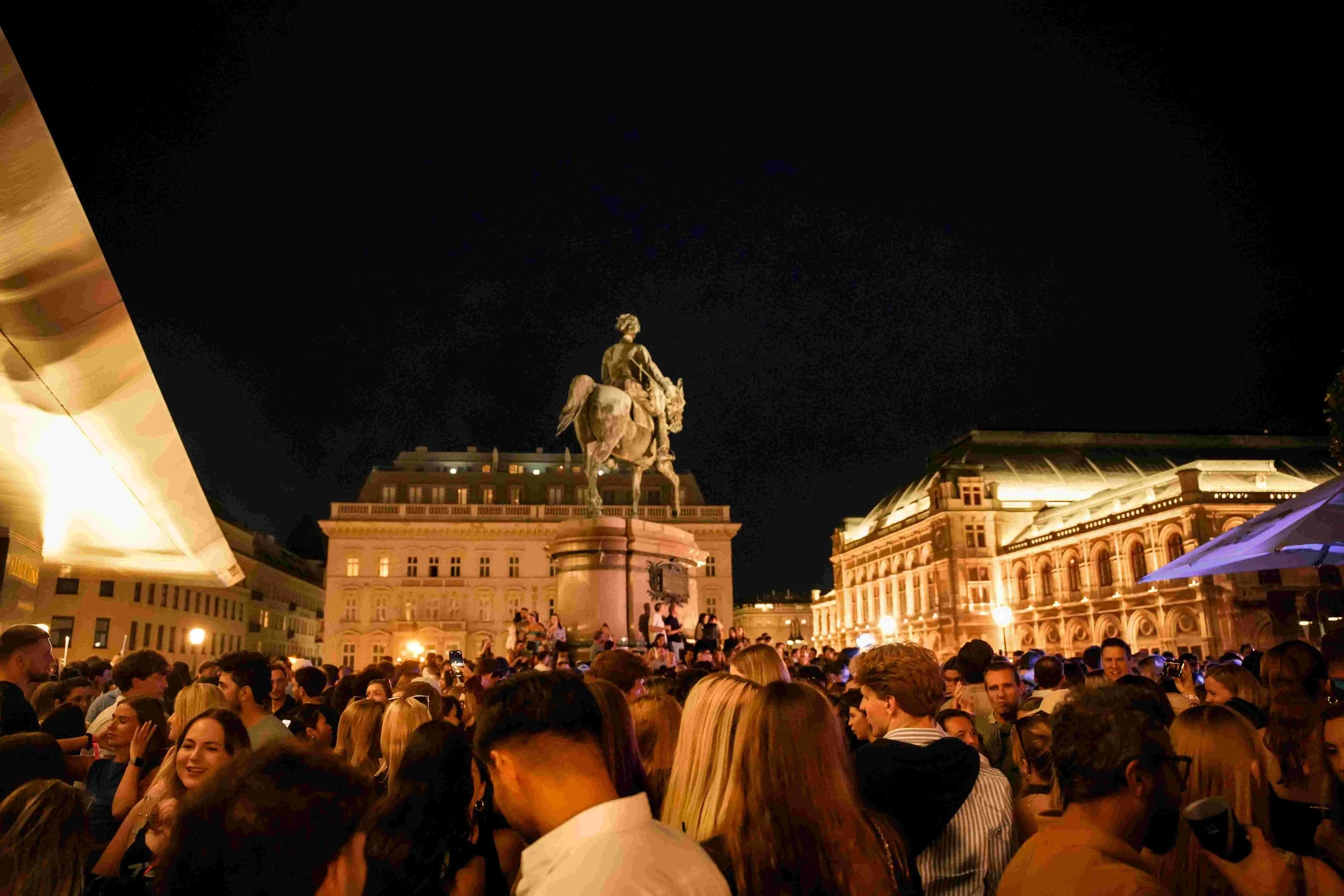 Crowd of people gathered outdoors at night near a historic building with a statue in the center, illuminated warmly.