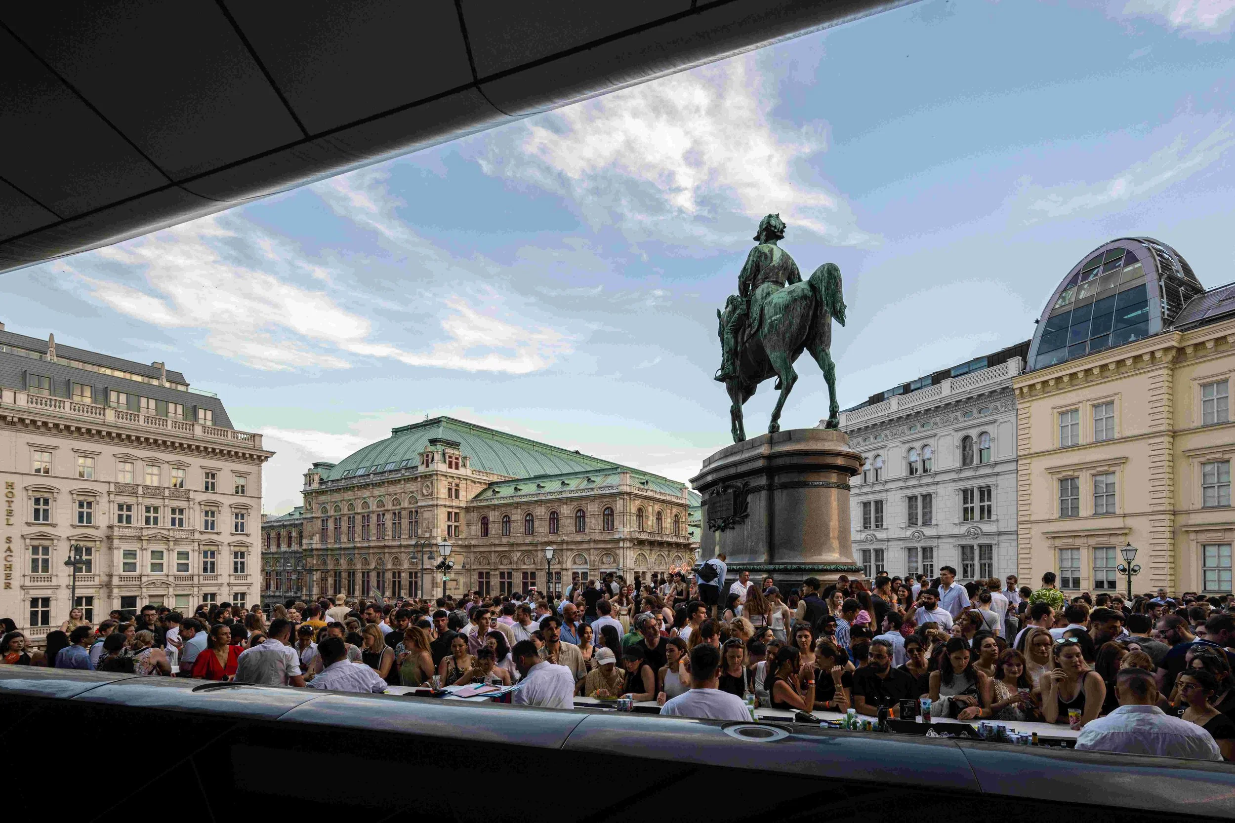 Crowd of people gathered in a plaza with historic buildings, a large statue of a man riding a horse, and a modern glass structure in the background, under a partly cloudy sky.