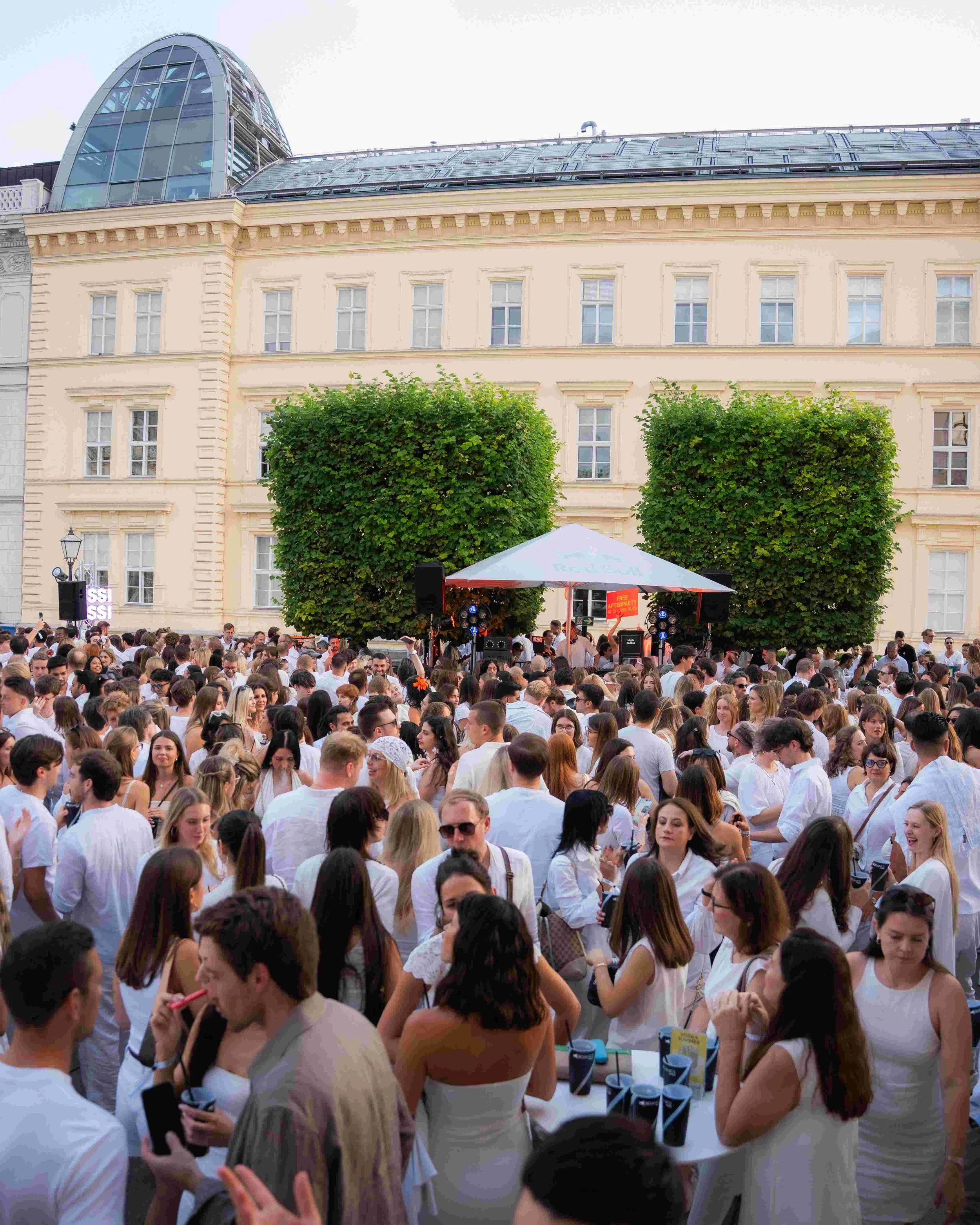 Crowd of people wearing white, gathered outdoors in front of a historic building with a modern glass roof structure, under a cloudy sky.