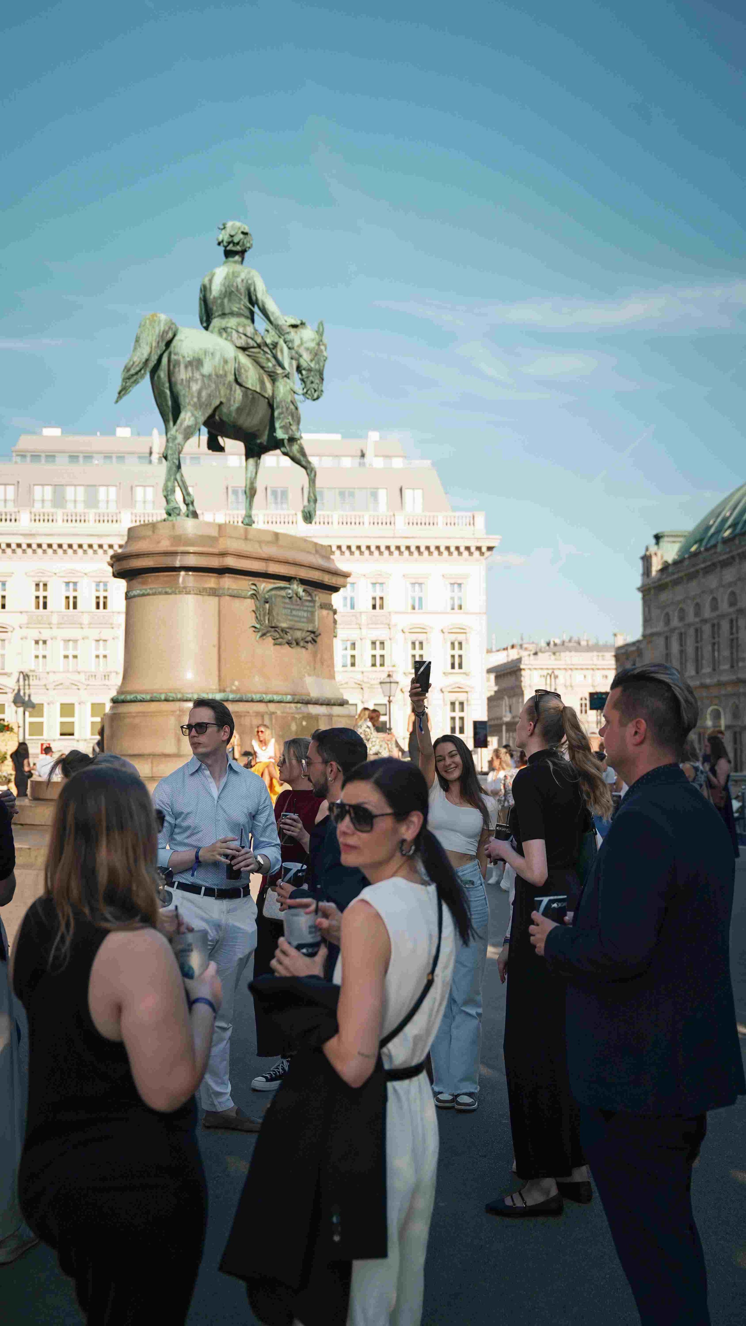 A group of people gathered in a city square with a large bronze statue of a man on a horse. The background features classical-style buildings and a clear blue sky.