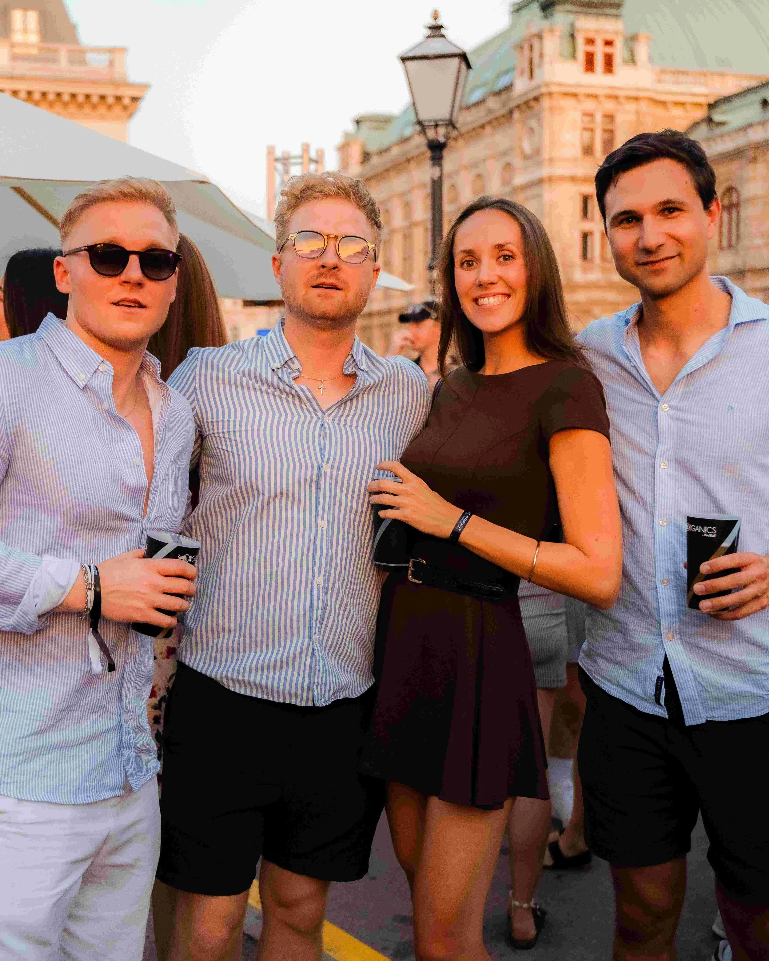Group of people standing outdoors at an event, with historic buildings and a streetlamp in the background, during sunset or late afternoon.