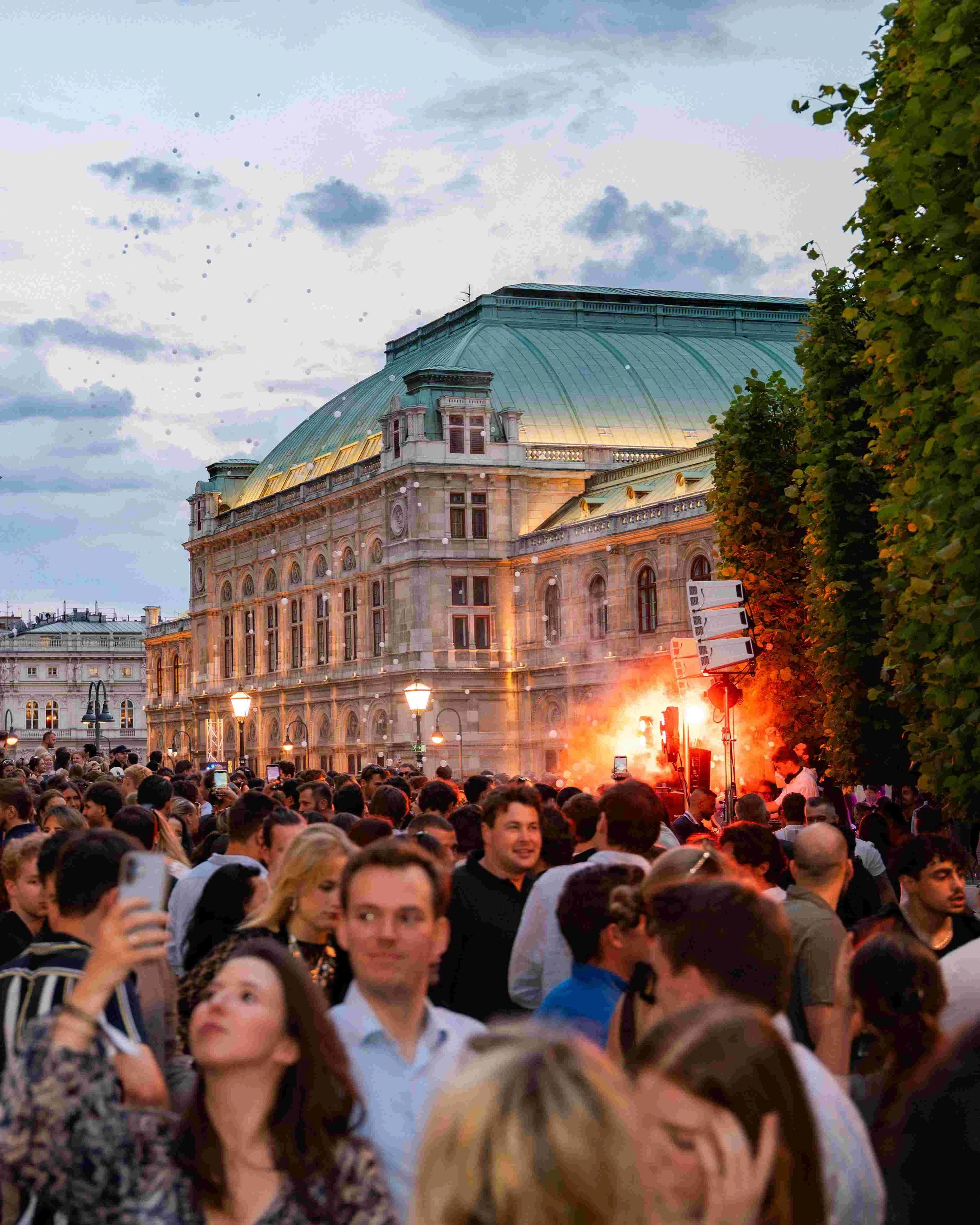 Crowd of people gathered outdoors during evening, with a historic building featuring a large domed roof and ornate architecture in the background, and a noticeable fire with bright flames and smoke among the crowd.