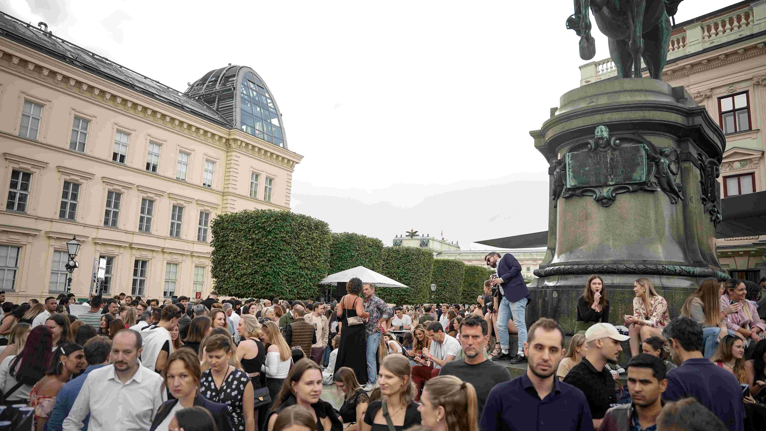 Large crowd of people gathered outdoors in front of historic and modern buildings, with a bronze equestrian statue on a tall pedestal to the right.