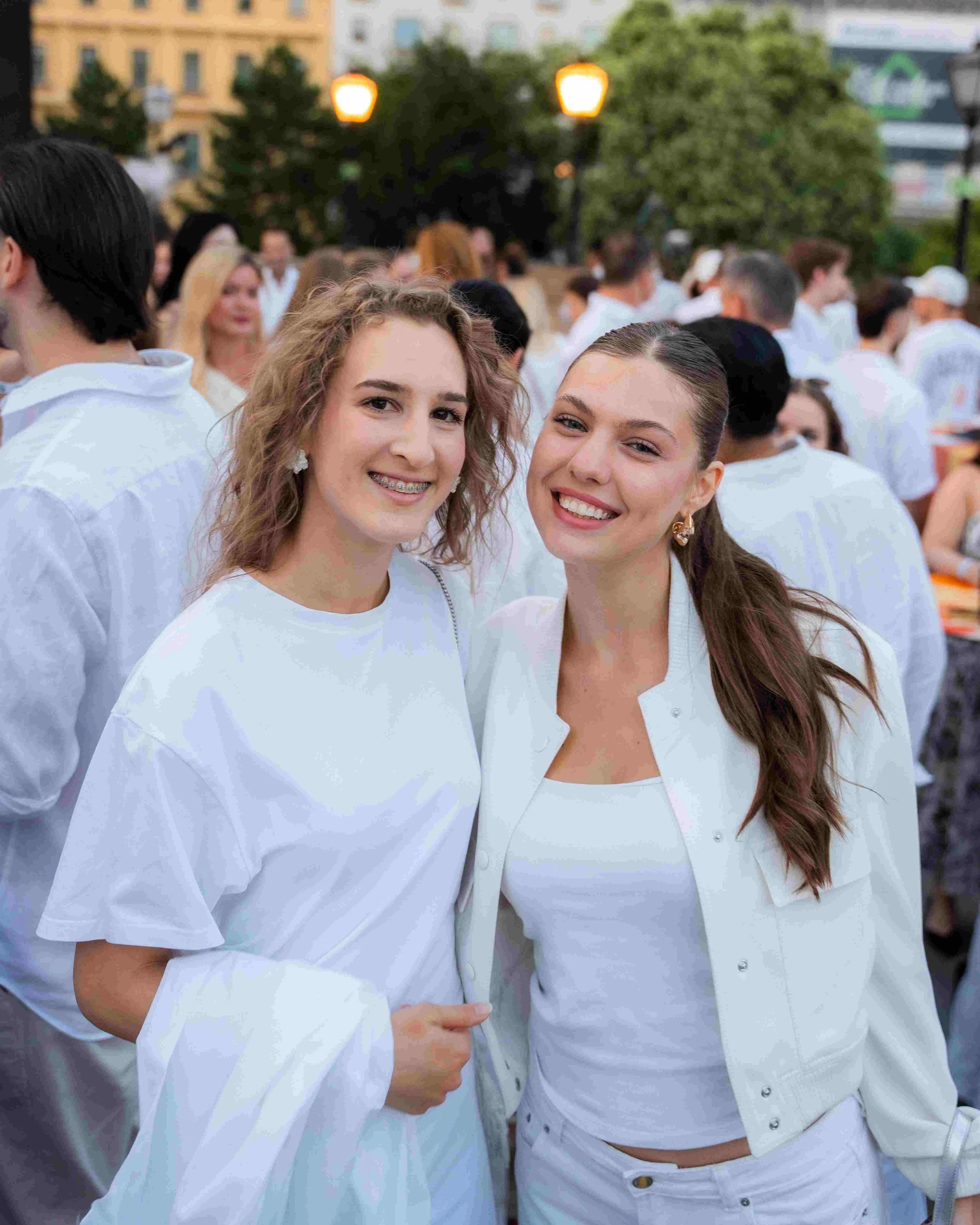 Smiling young women at an outdoor event, wearing white clothes, with blurred crowd and streetlights in the background.