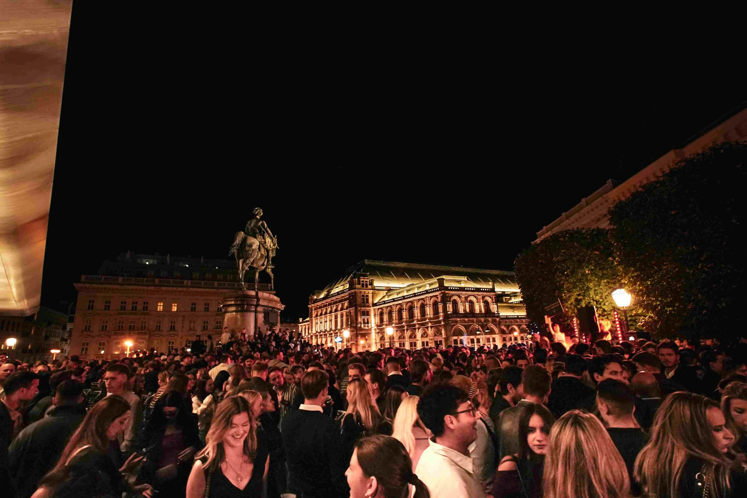 A large crowd gathered in an outdoor plaza at night, with many people standing and some sitting. In the background, an equestrian statue and illuminated historic buildings are visible. The scene is lively with warm lighting.