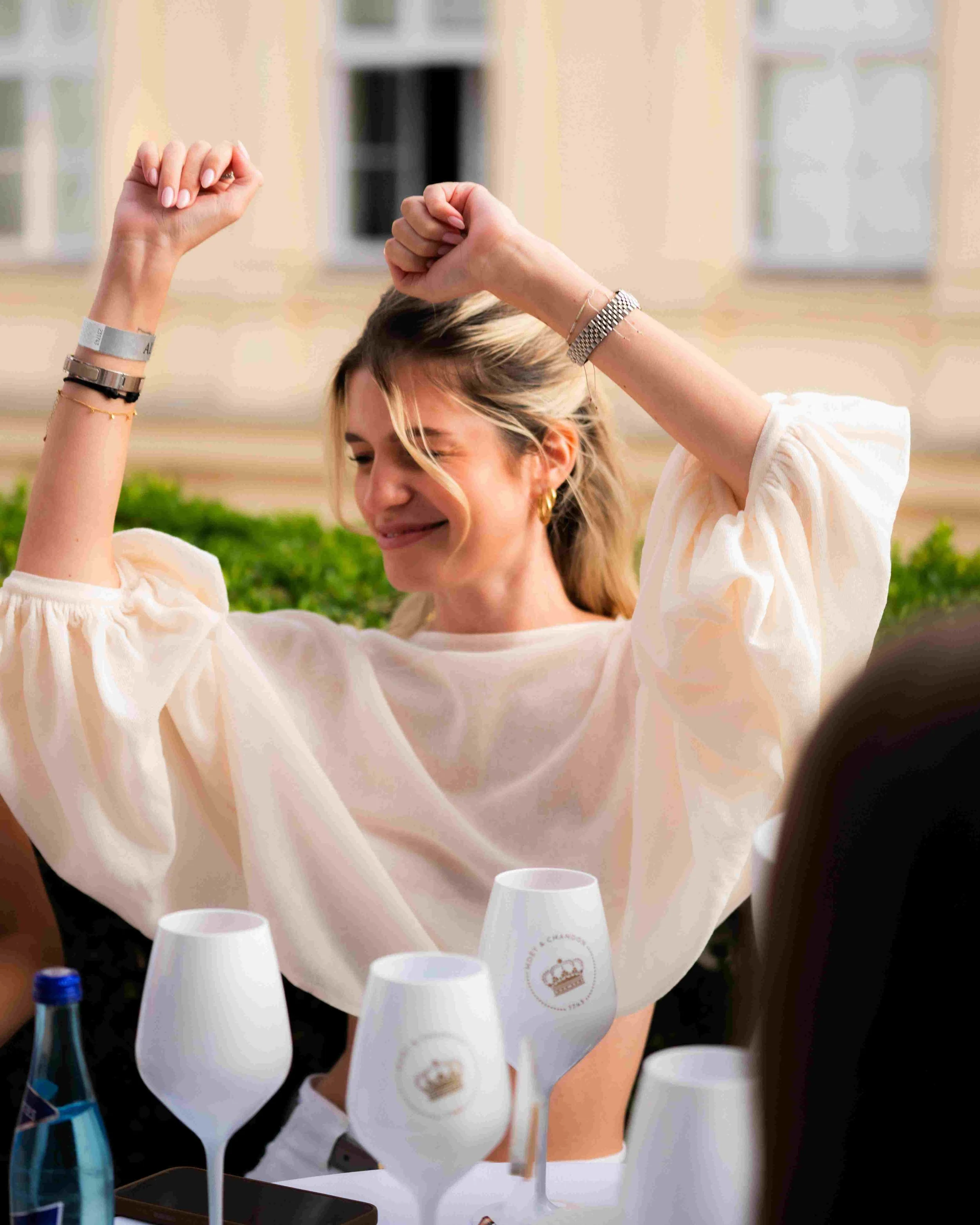 A woman with blonde hair smiling and raising her arms, wearing a white blouse, surrounded by white plastic cups and bottles on a table outdoors during daytime.