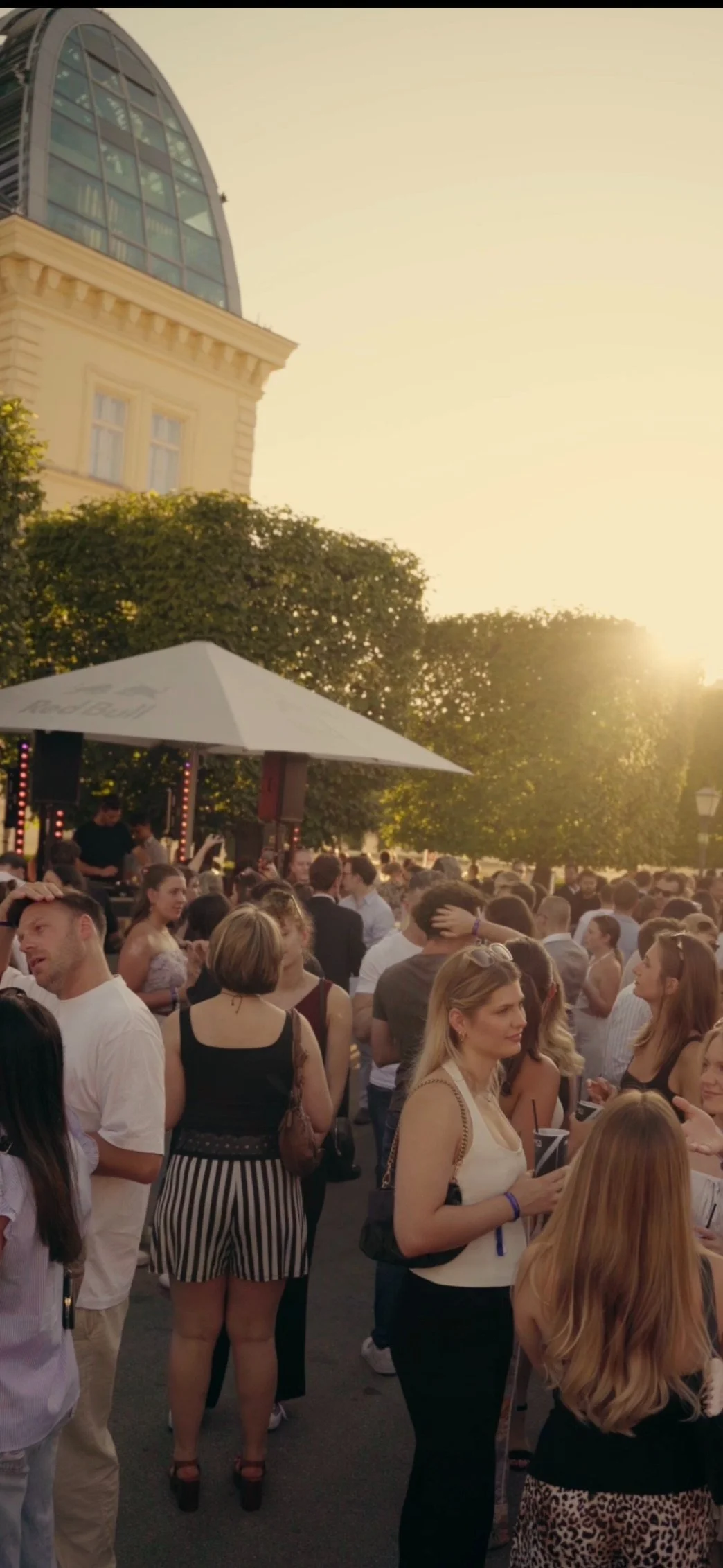 A crowded outdoor festival or concert during sunset with many people socializing, some with drinks, under a large white umbrella and trees.