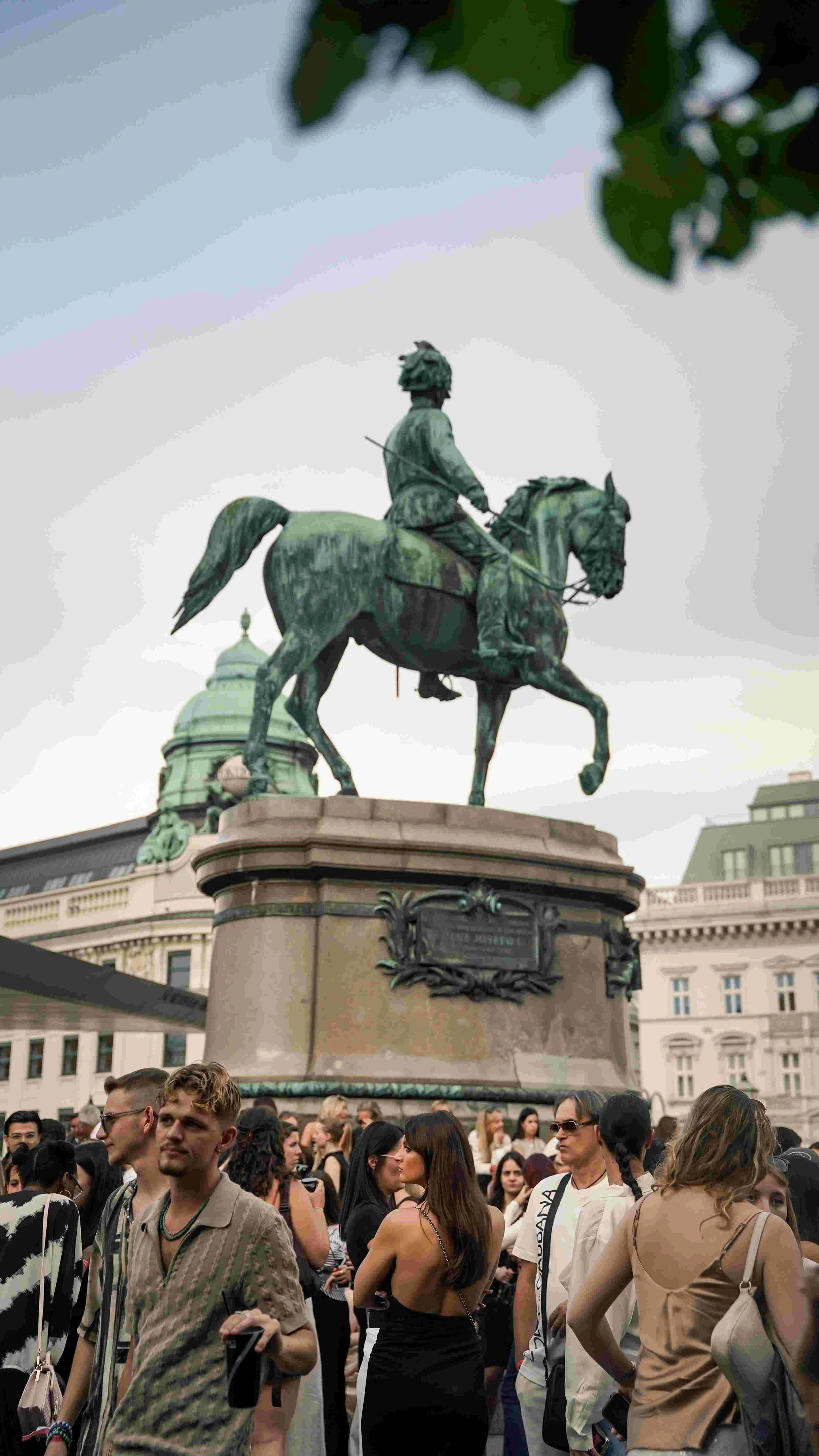 A crowd of people gathered beneath an equestrian statue of a man riding a horse, located in a city square with historic buildings and a cloudy sky in the background.