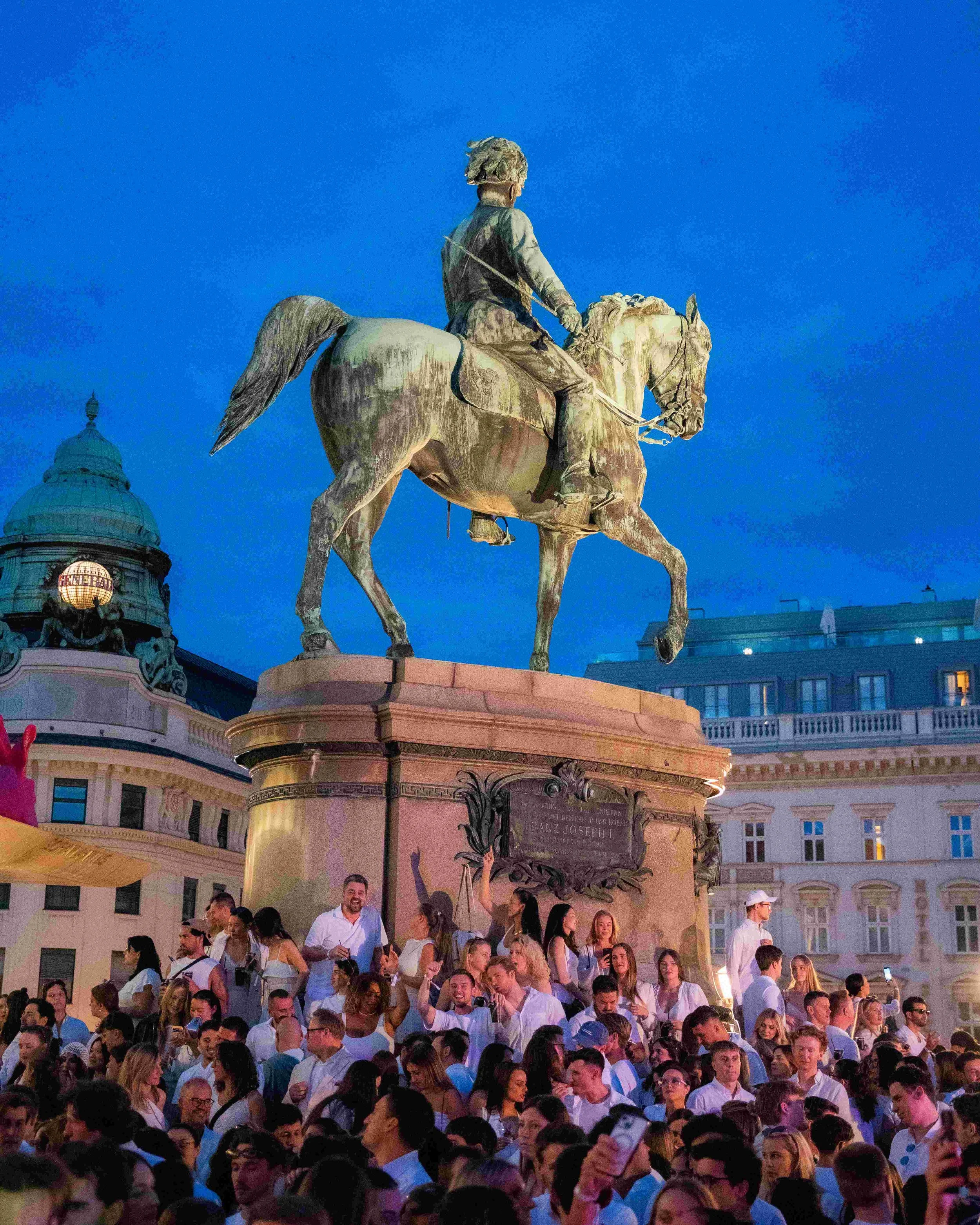 A large crowd of people gathered in front of an equestrian statue during the evening, with a historic building in the background and a blue sky overhead.
