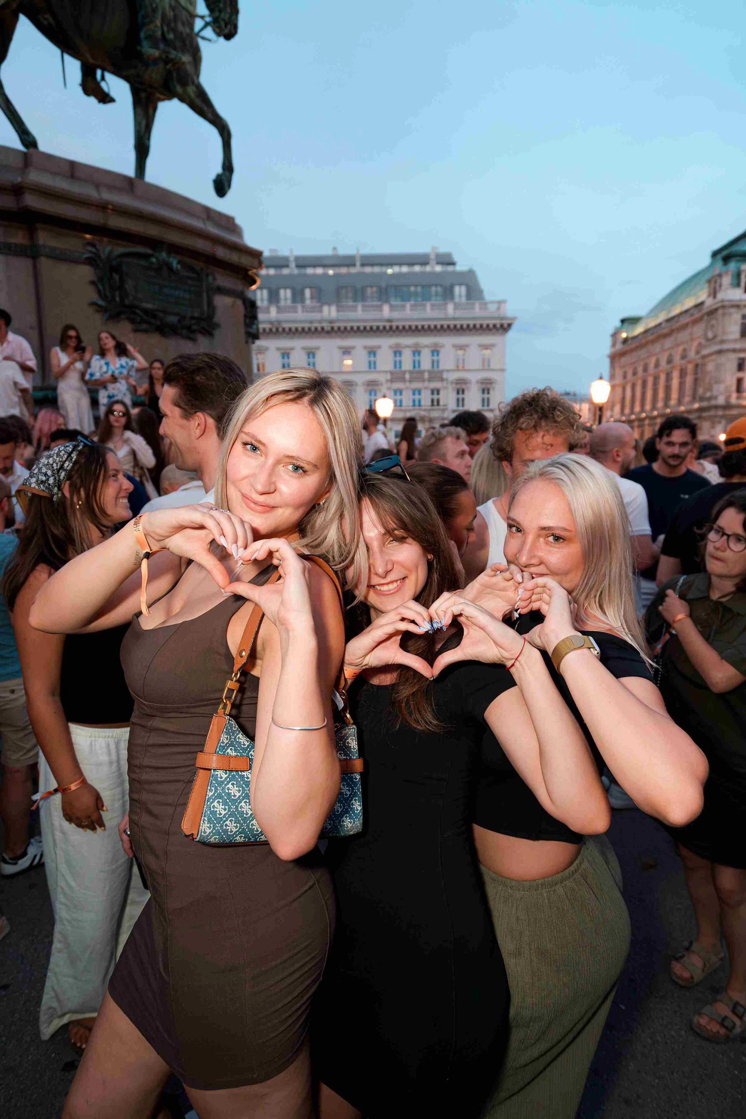 A group of young women making heart shapes with their hands during a crowded outdoor event at dusk in an urban setting.