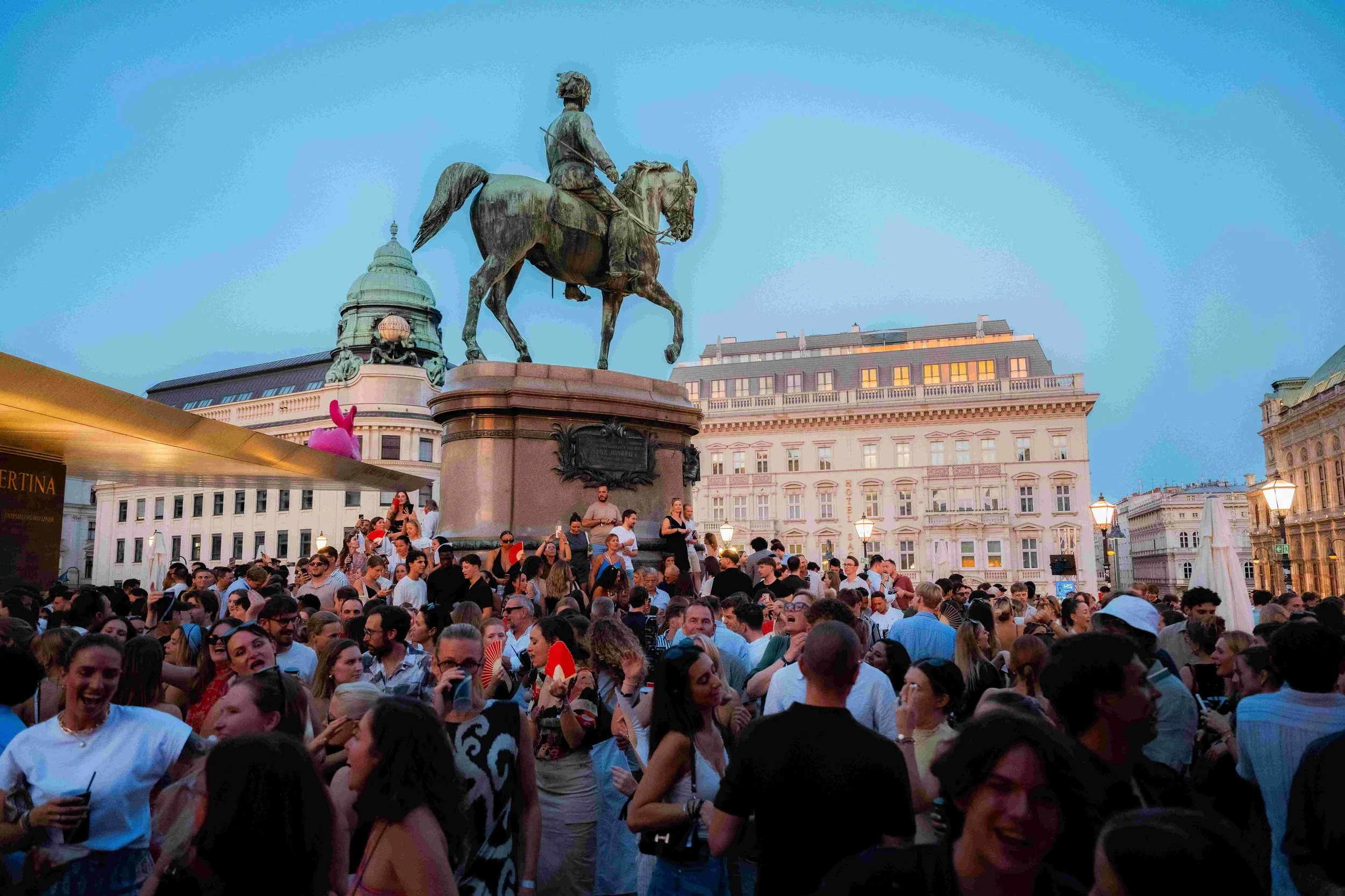 A large crowd of people gathered in a city square under a clear evening sky, with an equestrian statue of a historical figure prominently displayed above them. Buildings with detailed architecture surround the square, and some people are holding fans
