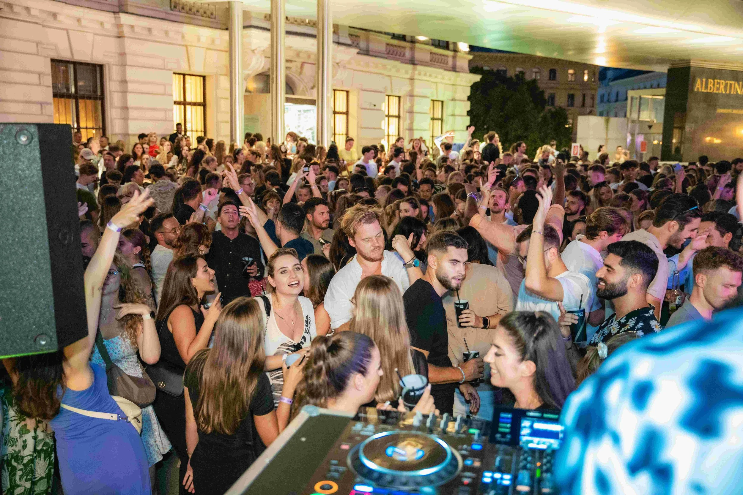 A large crowd of young people at a nighttime outdoor event, dancing and socializing behind DJ equipment on a platform.