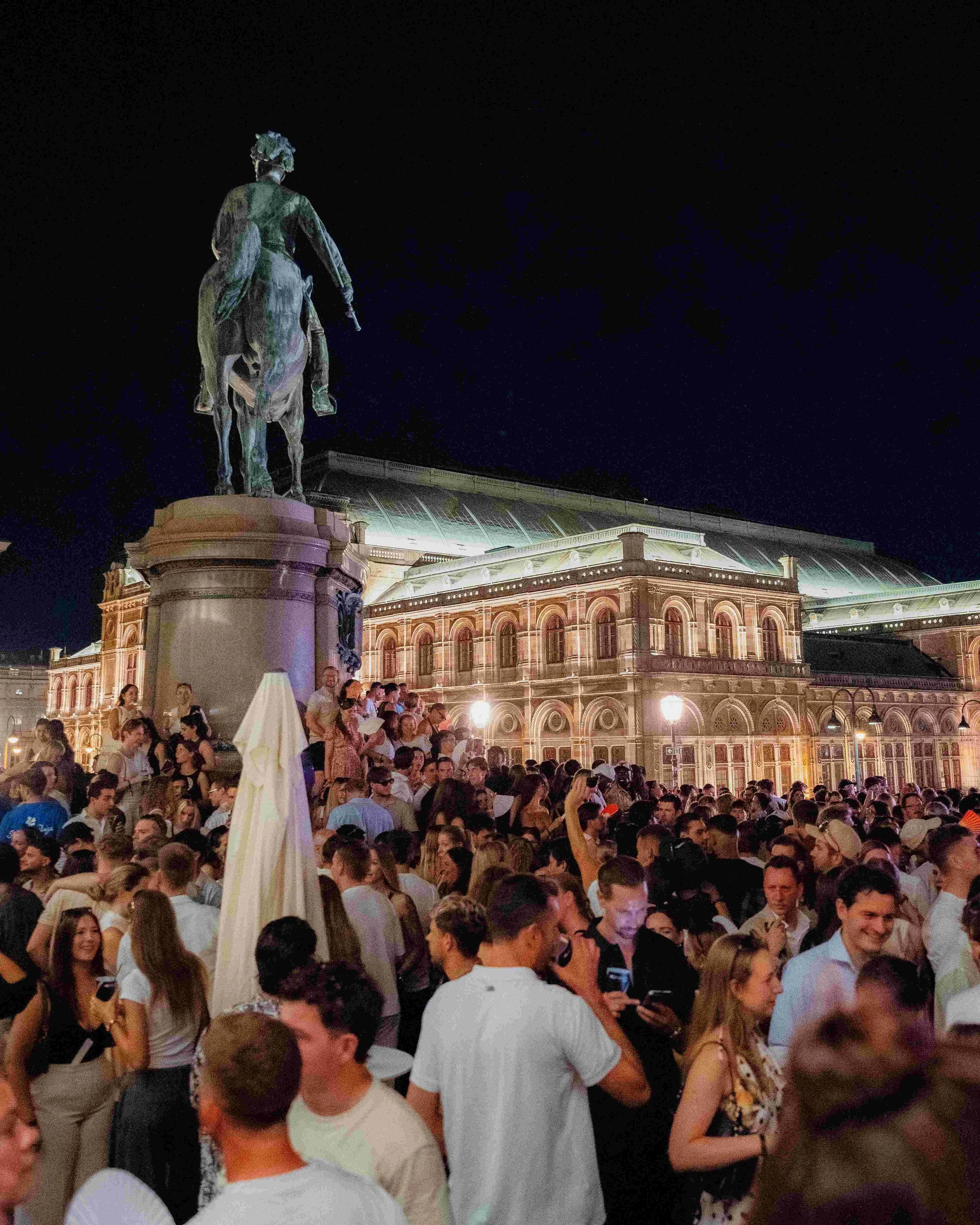 A large crowd of people gathered outdoors at night, with a historic building illuminated in the background. There is a statue of a mounted figure on a pedestal in the foreground.