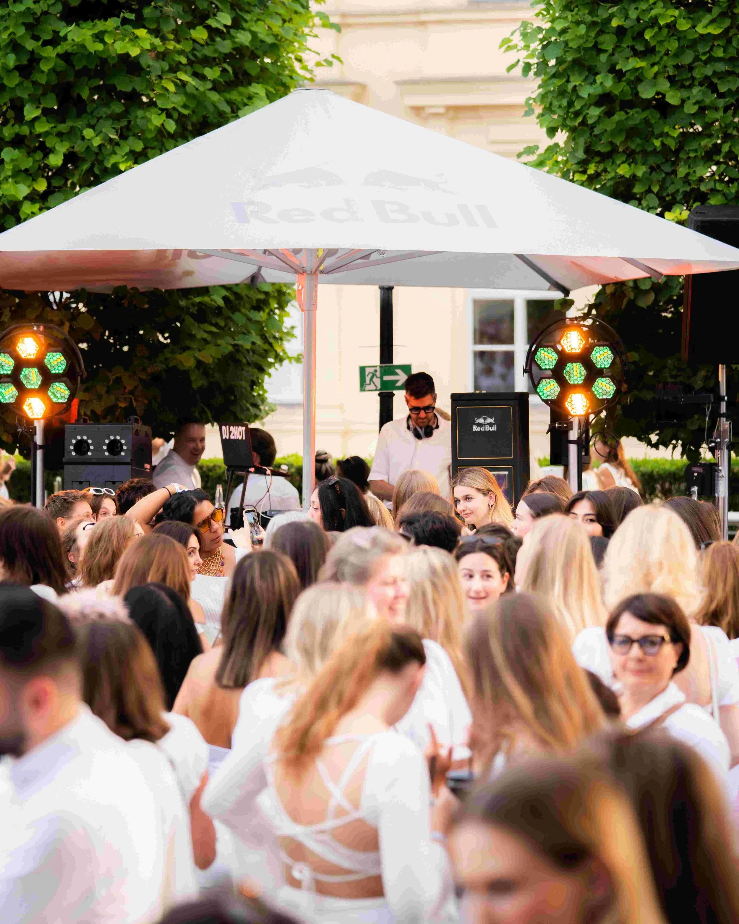 A crowd of people gathered outdoors under a white Red Bull-branded umbrella, with a DJ playing music behind them. There are bright stage lights and a building with a window and trees in the background.
