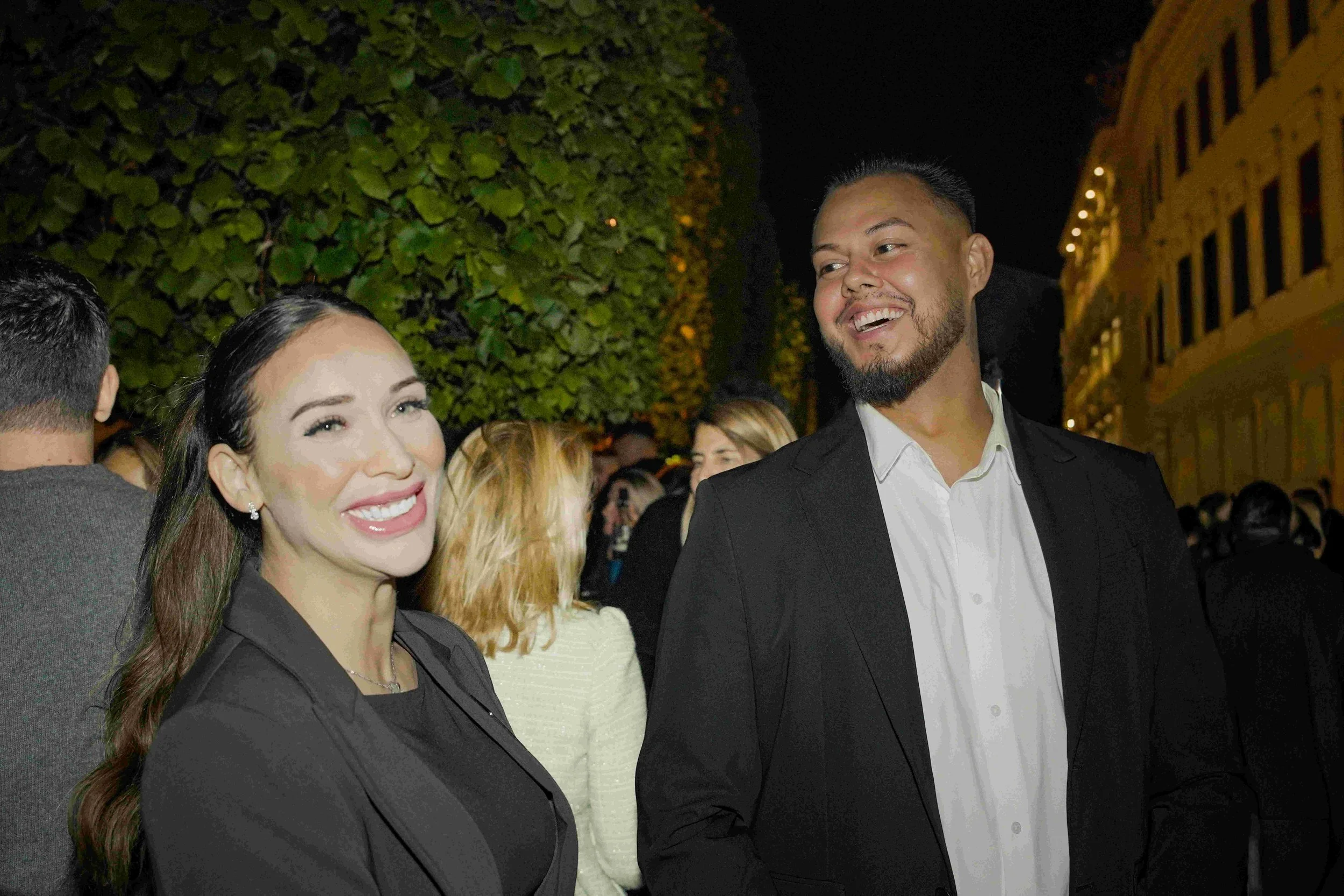A group of people at an outdoor nighttime event, smiling and dressed in formal attire, with a woman with dark hair, earrings, and a black blazer in the foreground and a man with a beard and short hair in a black suit and white shirt beside her. There