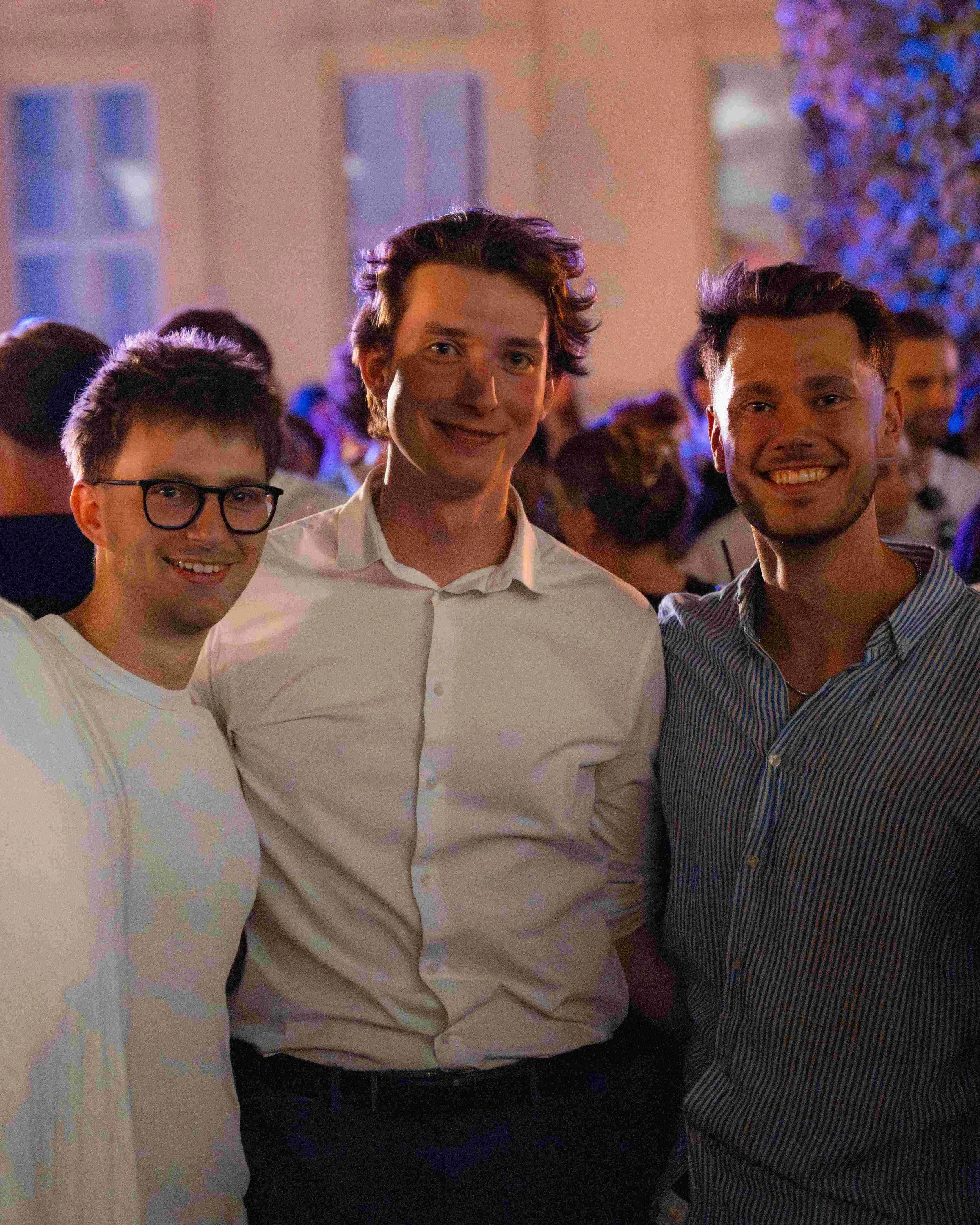 Three young men are standing together at a social event, smiling and wearing casual clothing, with a crowd of people in the background.