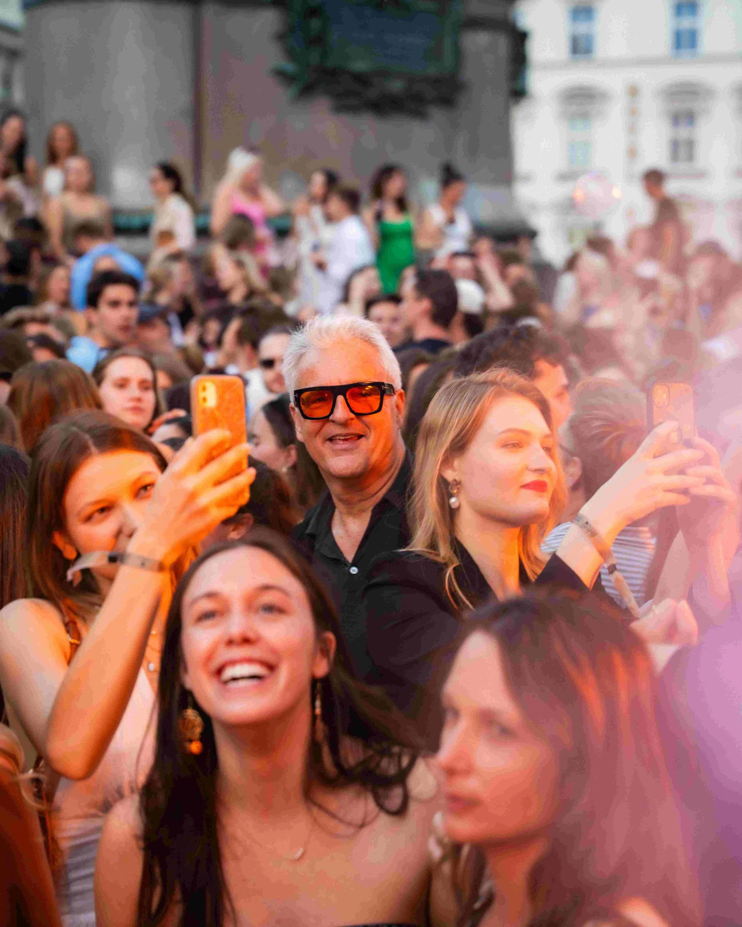 Crowd of people gathered outdoors, with many taking photos or videos, some smiling and others talking. There's a man with white hair and sunglasses in the center, and a woman with long hair in front.