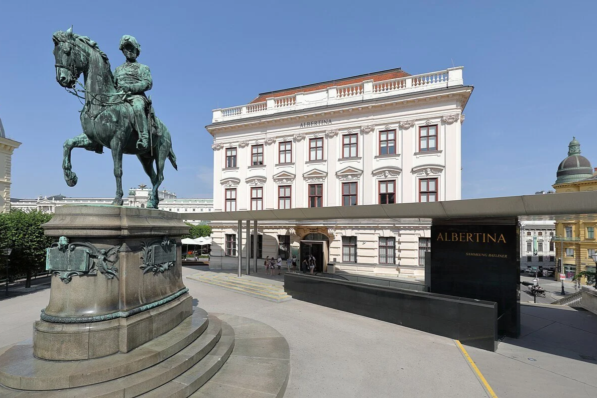 View of an ornate white building with a sign reading "Albertina" in Vienna, Austria. In front of the building, there is a large bronze statue of a man in a military uniform on horseback with detailed decorations at the base. People are entering and exiting the building, and the sky is clear and sunny.