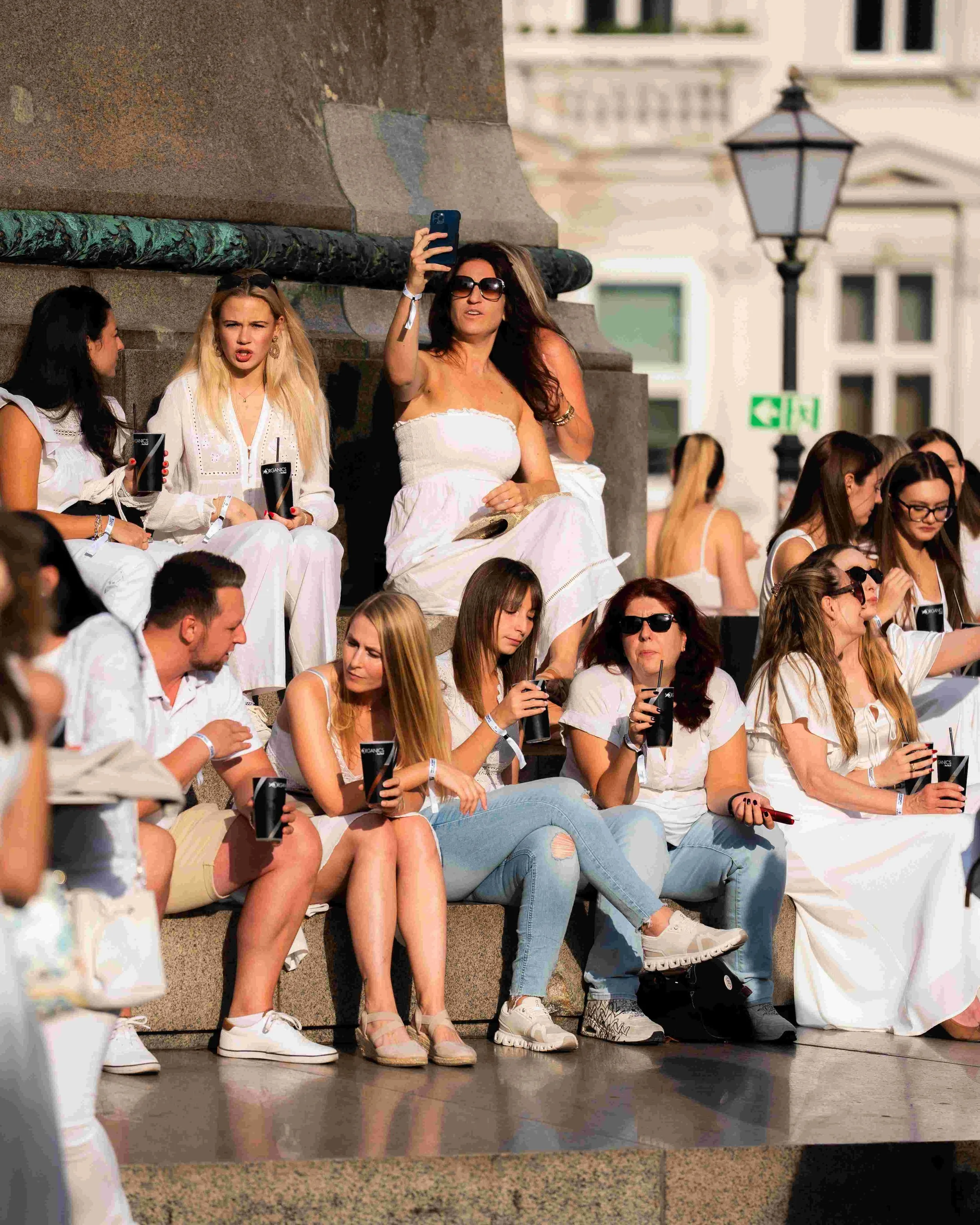 A group of women sitting on stone steps outdoors, some holding drinks, with three women in the foreground wearing white outfits. One woman in sunglasses is taking a selfie. Other women are engaged in conversation or looking at their phones. The setti