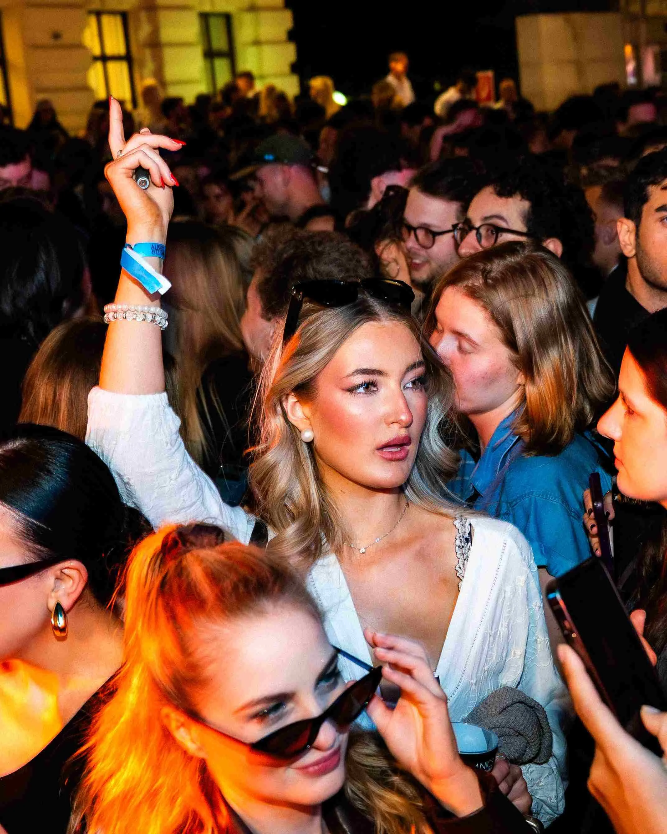 Crowd of people at a social event with a woman in the foreground wearing sunglasses on her head, pearl earrings, and a white lace top, surrounded by other attendees, some wearing glasses and colorful clothing.
