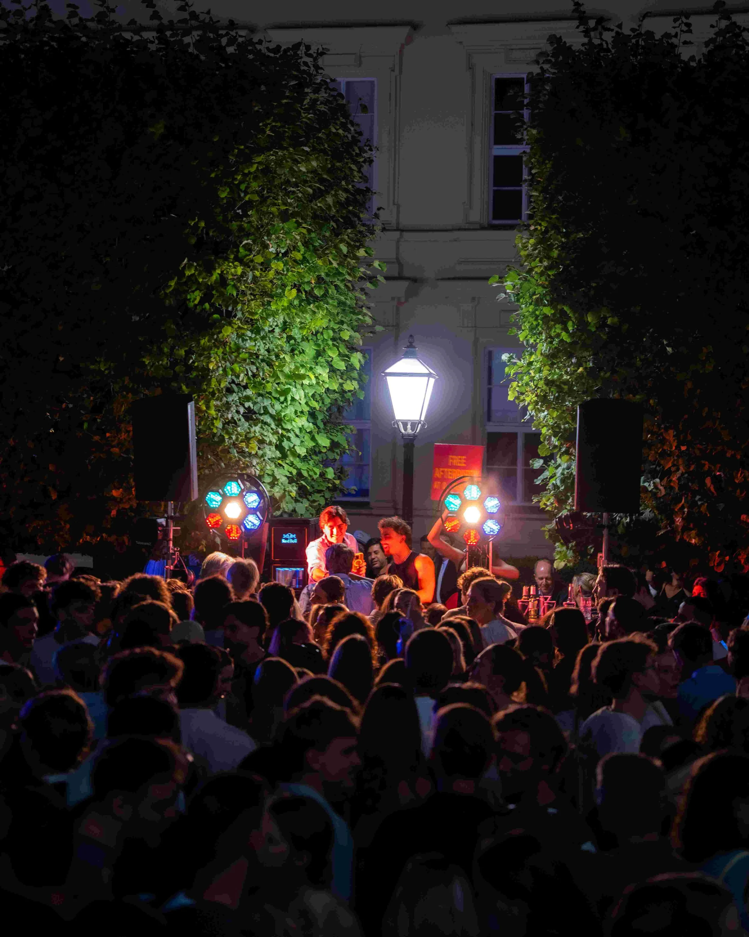 Nighttime outdoor scene with a large crowd gathered in front of a stage, surrounded by trees. Bright stage lights and a person speaking or performing are visible on the stage. A street lamp is also illuminated in the background.