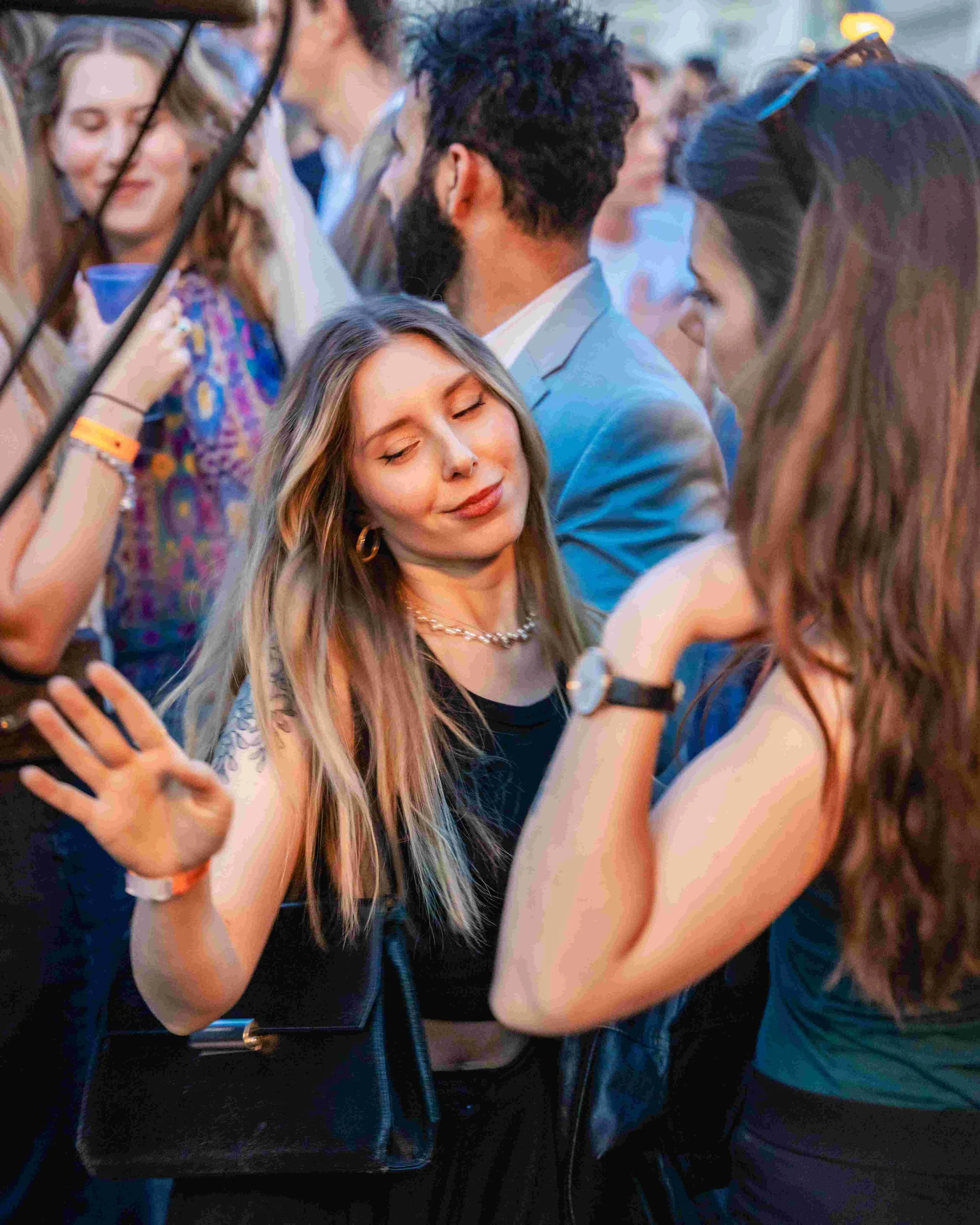 Group of young people at a social gathering, some taking selfies, smiling, and enjoying each other's company in a lively outdoor setting.