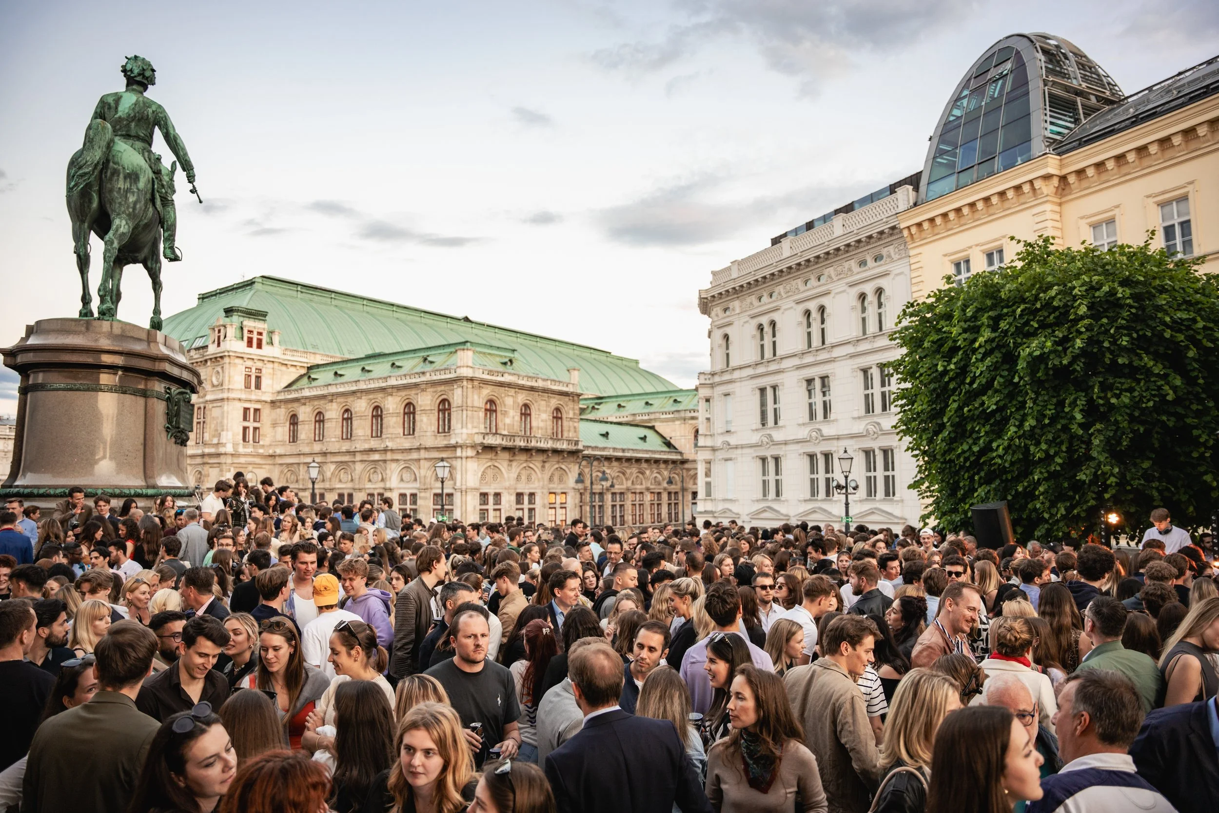 Crowd of people gathered outdoors in front of historic and modern buildings under a cloudy sky.