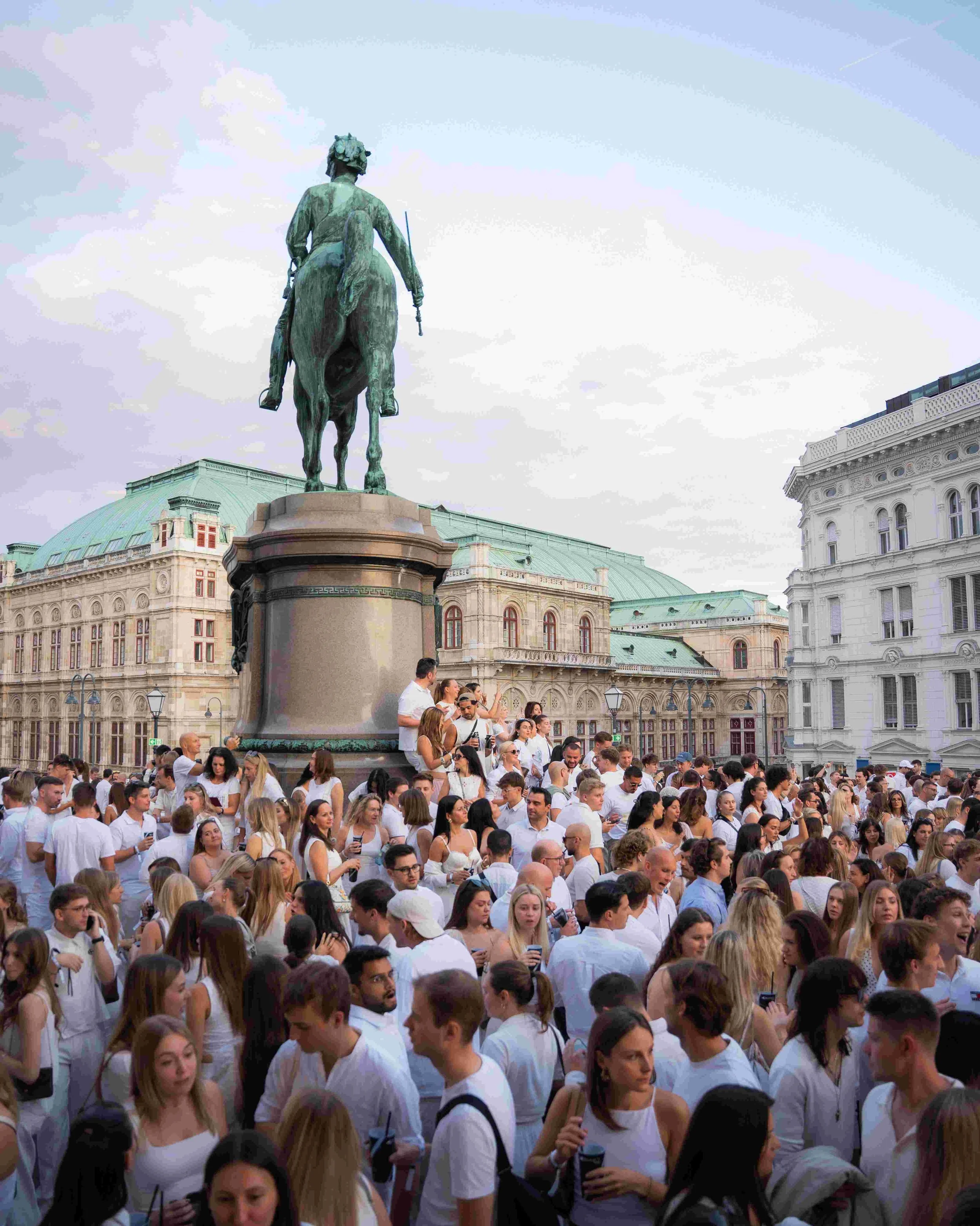A large crowd of people dressed in white gathered in front of a historic building with a statue of a mounted figure on a pedestal in the background.
