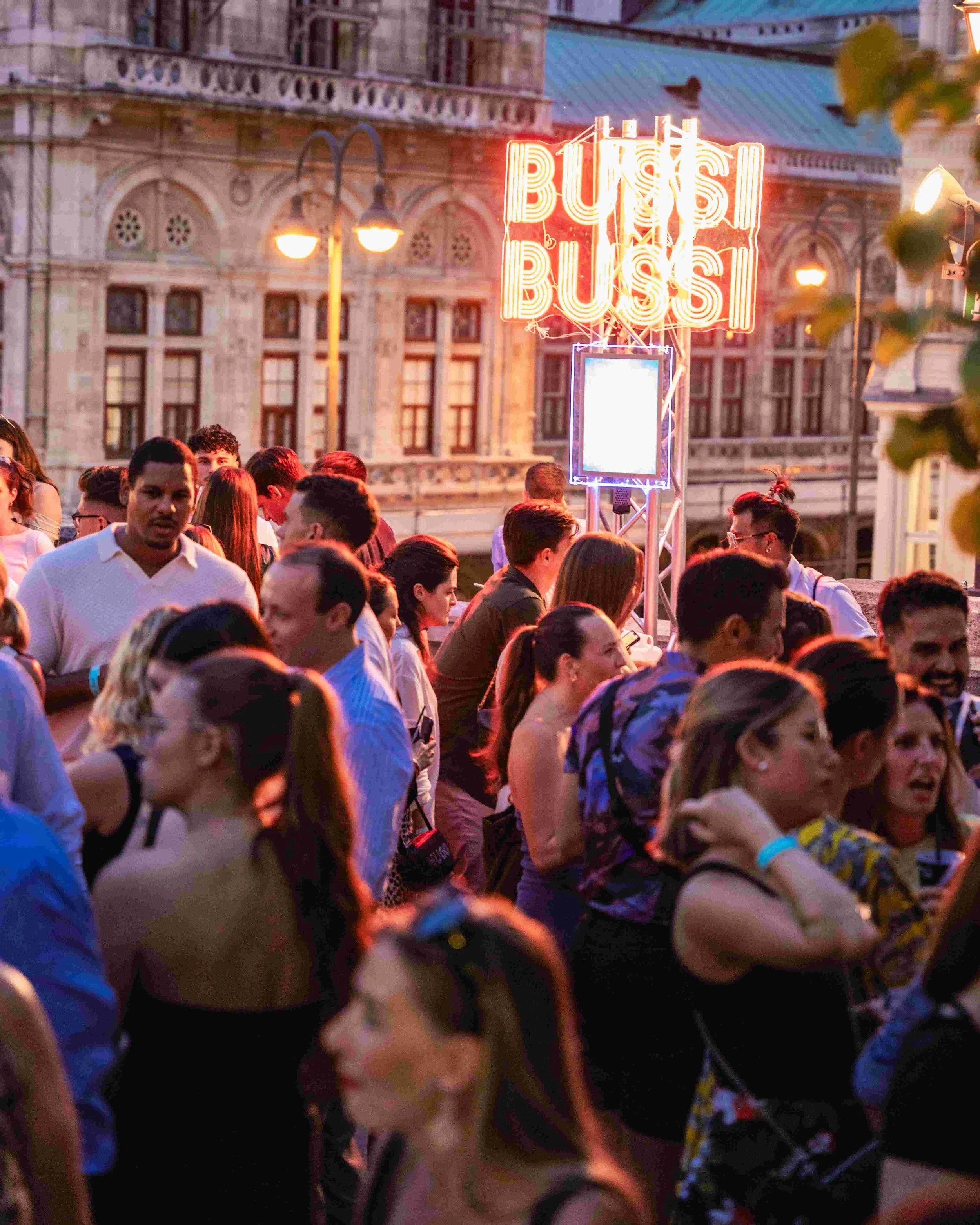 A crowd of people gathered outdoors during twilight, with some people smiling and others looking around; a brightly lit neon sign reads 'BUSY BUSY', and there are historic buildings with intricate architecture in the background.