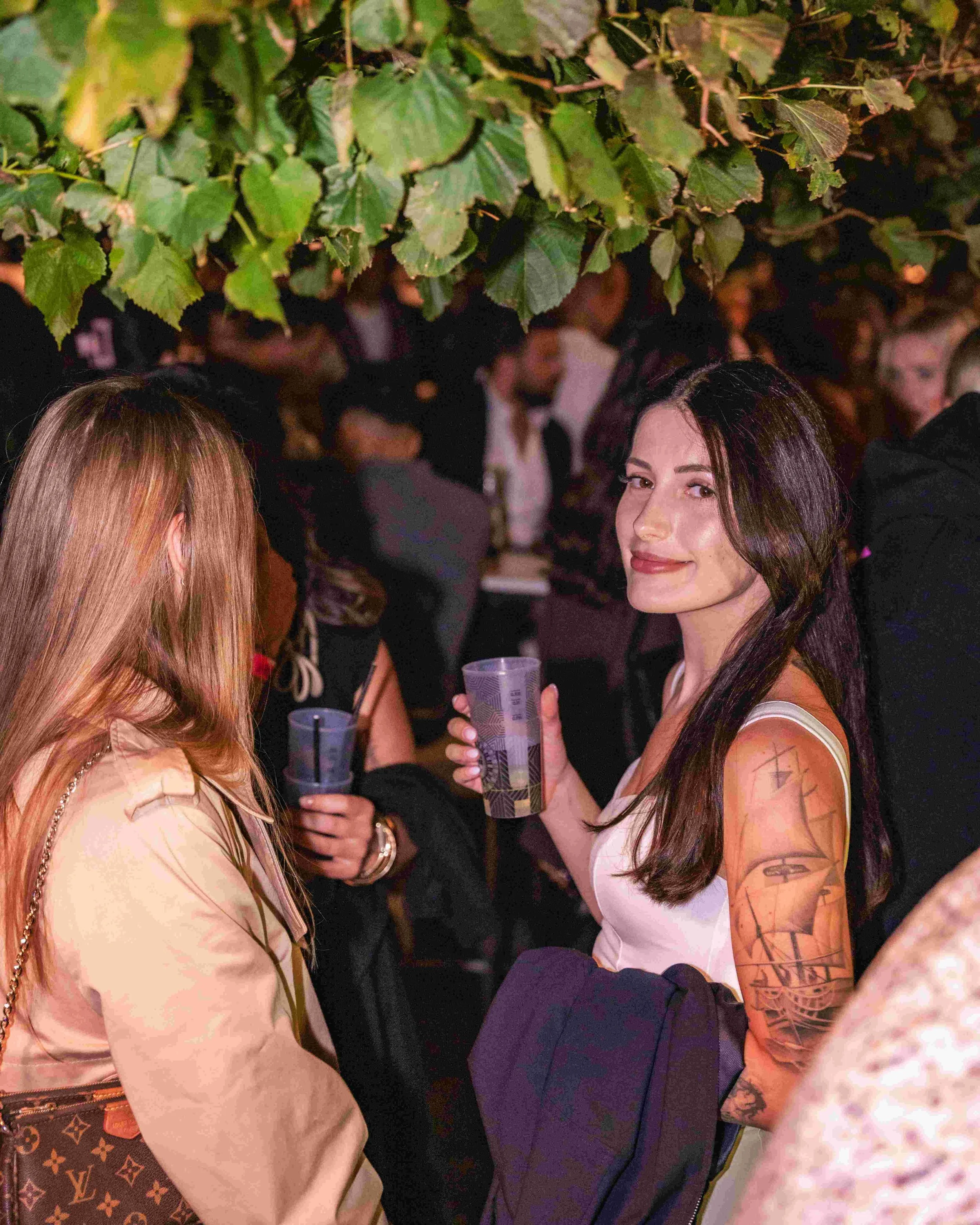 A group of people socializing at a party or social gathering with green leaves hanging overhead. The focus is on a woman with long dark hair and tattoos on her arm, holding a drink and smiling at the camera. Other people are visible in the background