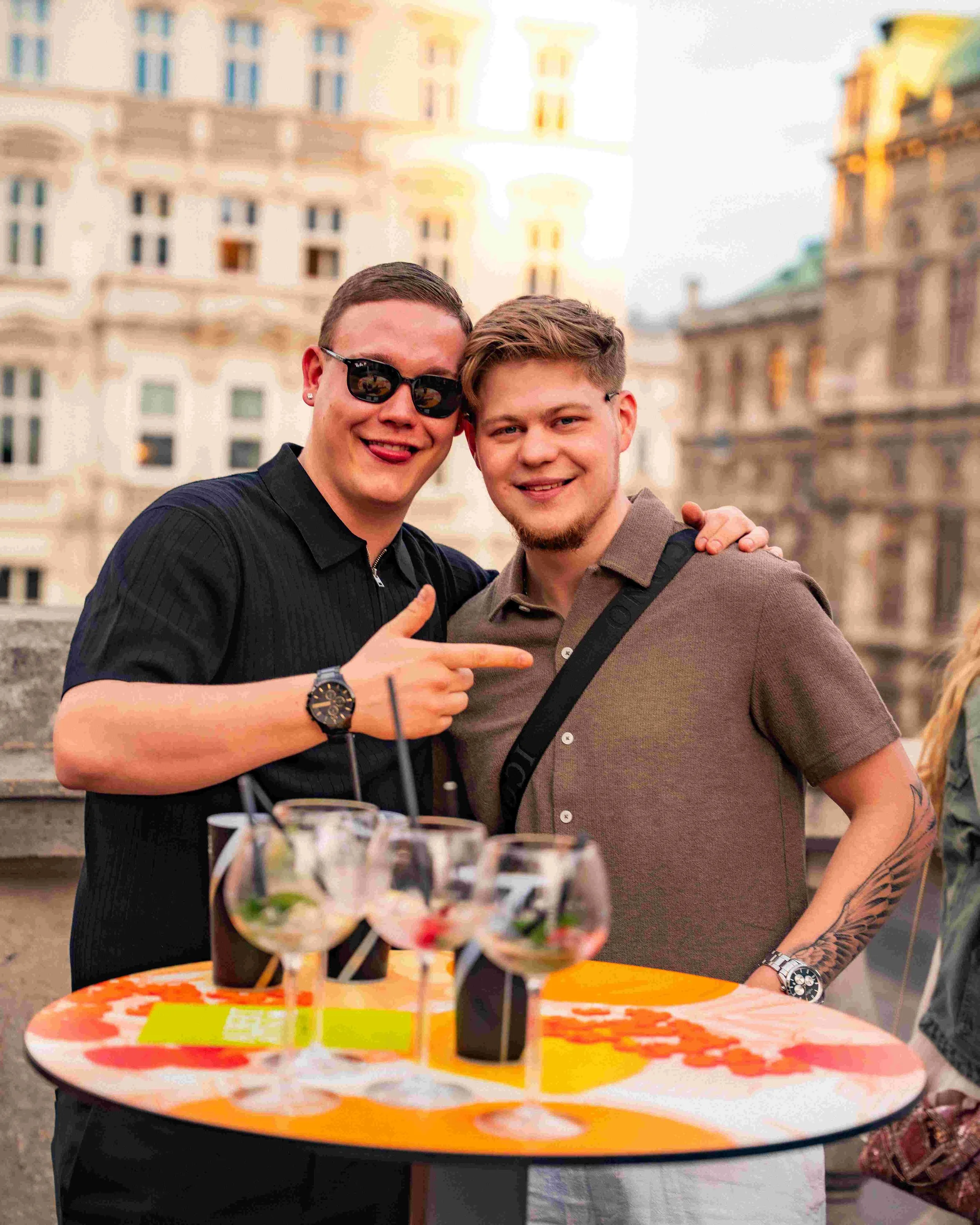 Two young men smiling, standing outdoors at sunset. One wearing sunglasses and a black shirt, the other with a beard and tattoos on his arm, wearing a brown shirt. A table with colorful drinks garnished with fruit is in the foreground, and buildings 