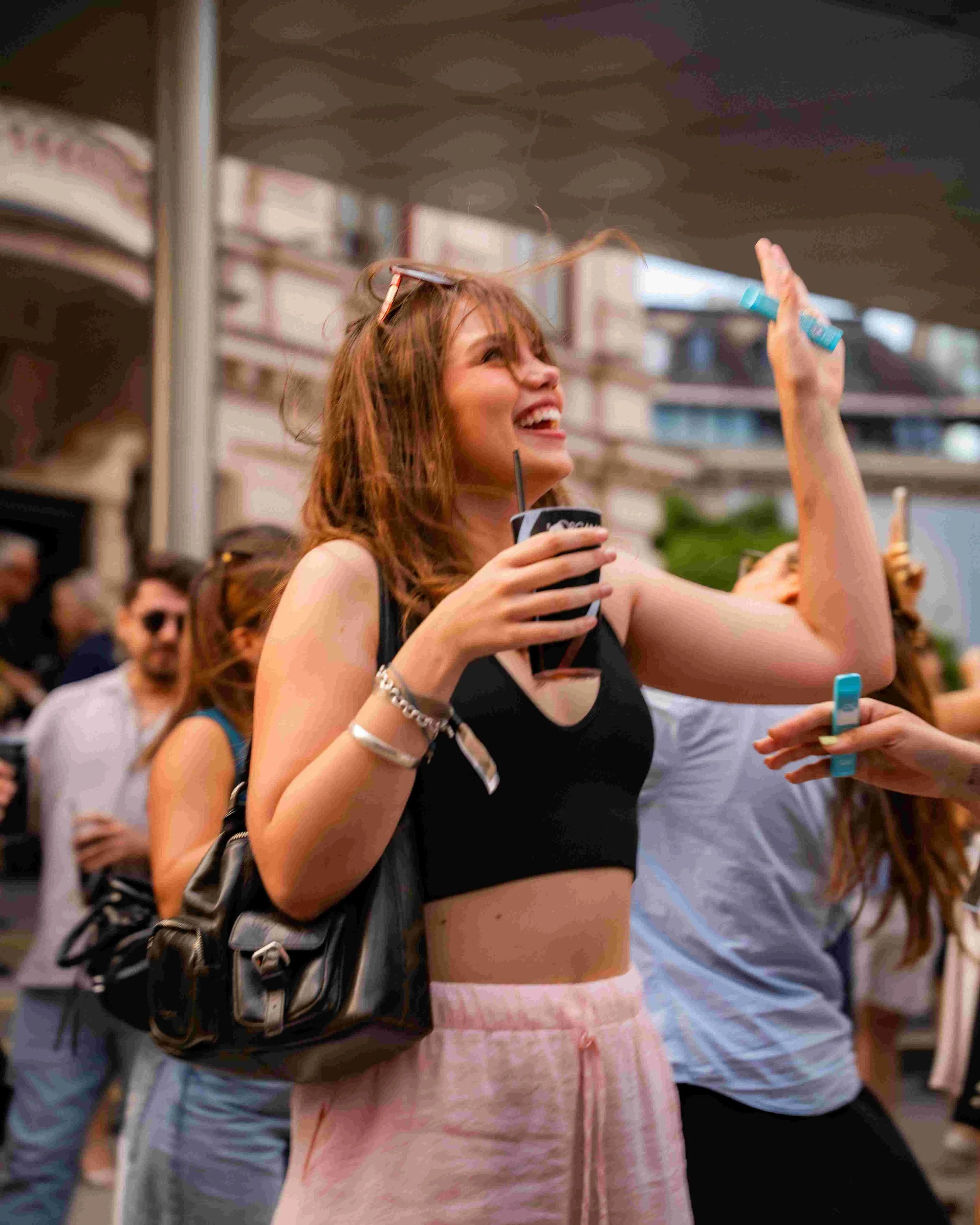 A young woman with long, red hair smiling and enjoying herself at a crowded outdoor event. She is holding a drink in one hand and a small blue object in the other, with sunglasses resting on her head. She is wearing a black tank top and pink shorts, 
