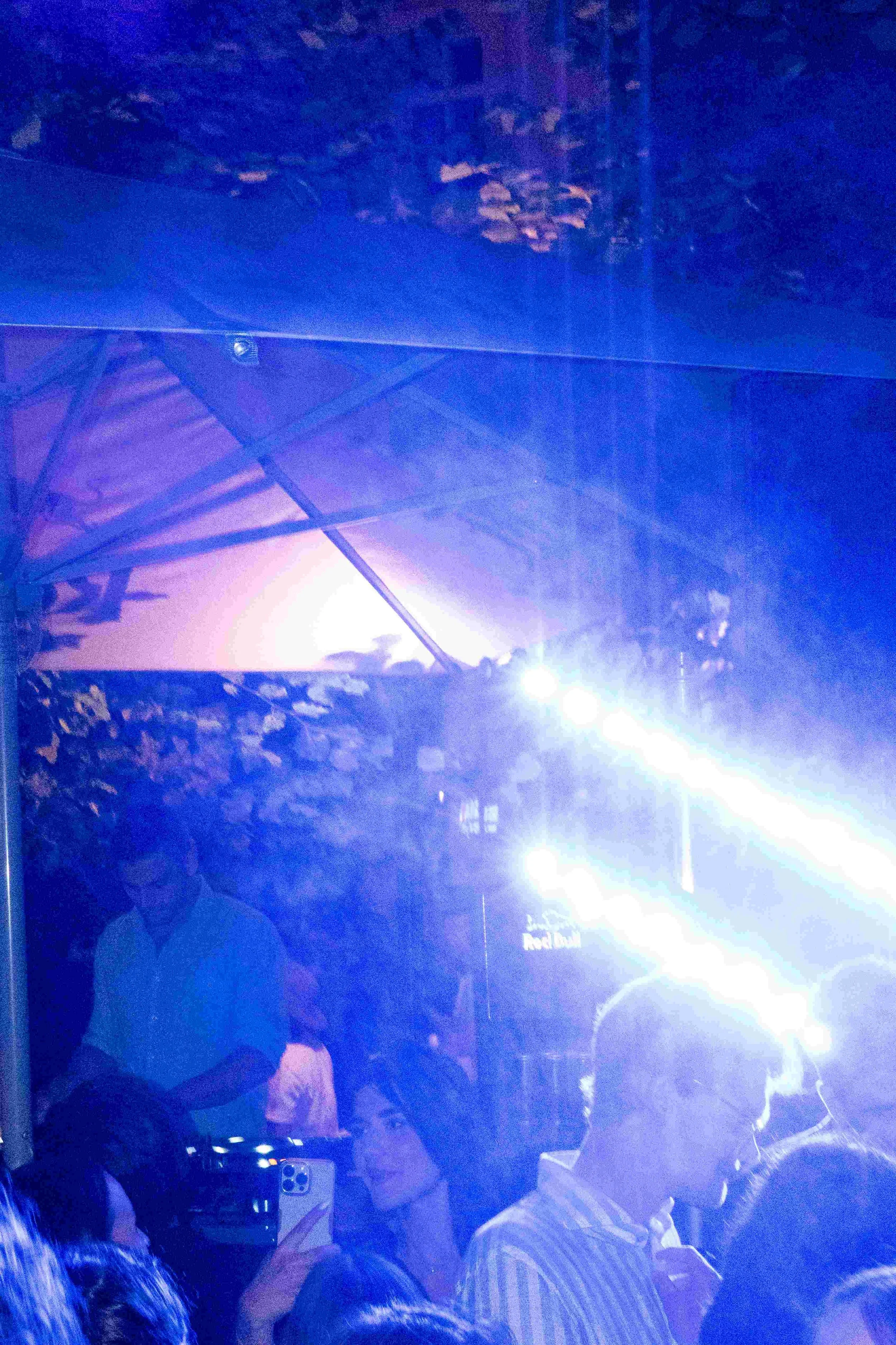 People gathered at an event or concert, illuminated by a bright blue and purple light, with some individuals holding phones.