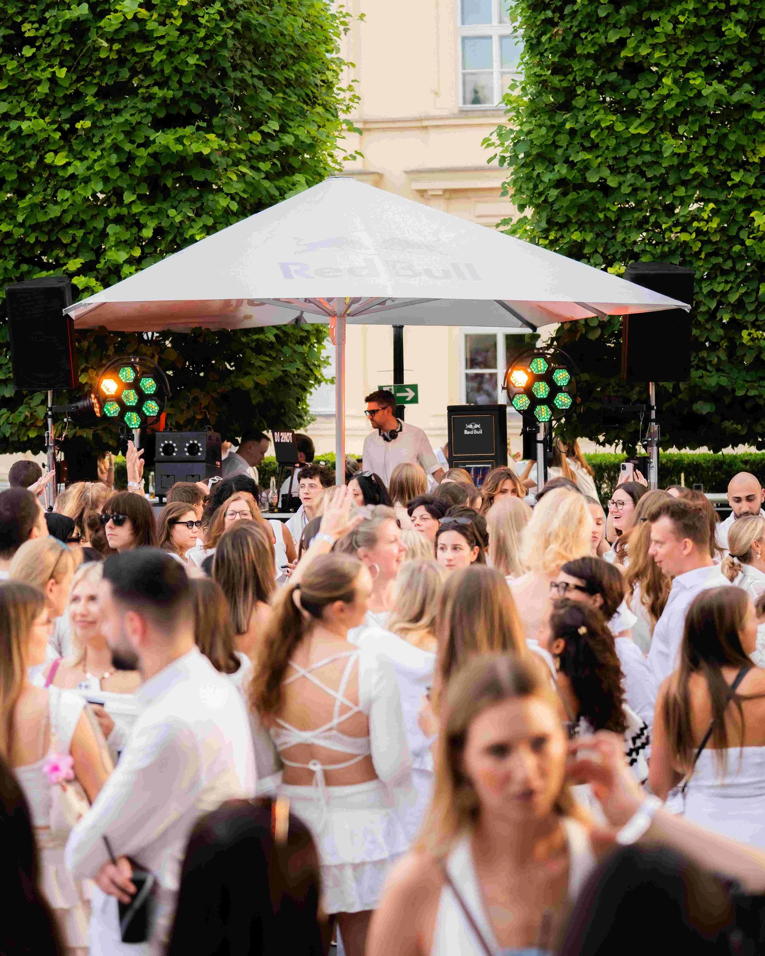 A crowded outdoor event with many people gathered under a white tent, with two green and orange stage lights and a DJ or performer wearing sunglasses and headphones in the center.