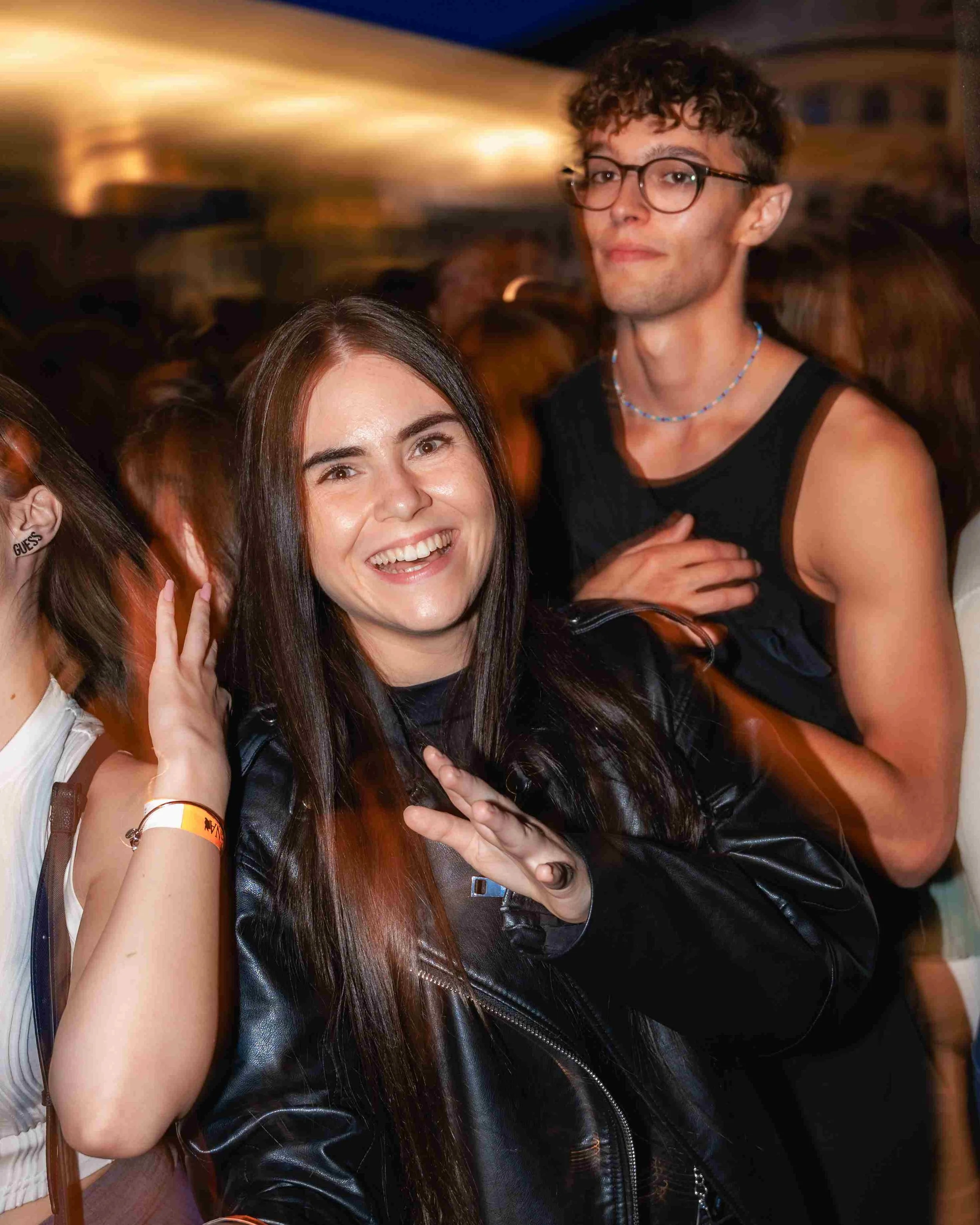 Two young women at a party smiling and posing for the camera with blurred people in the background.