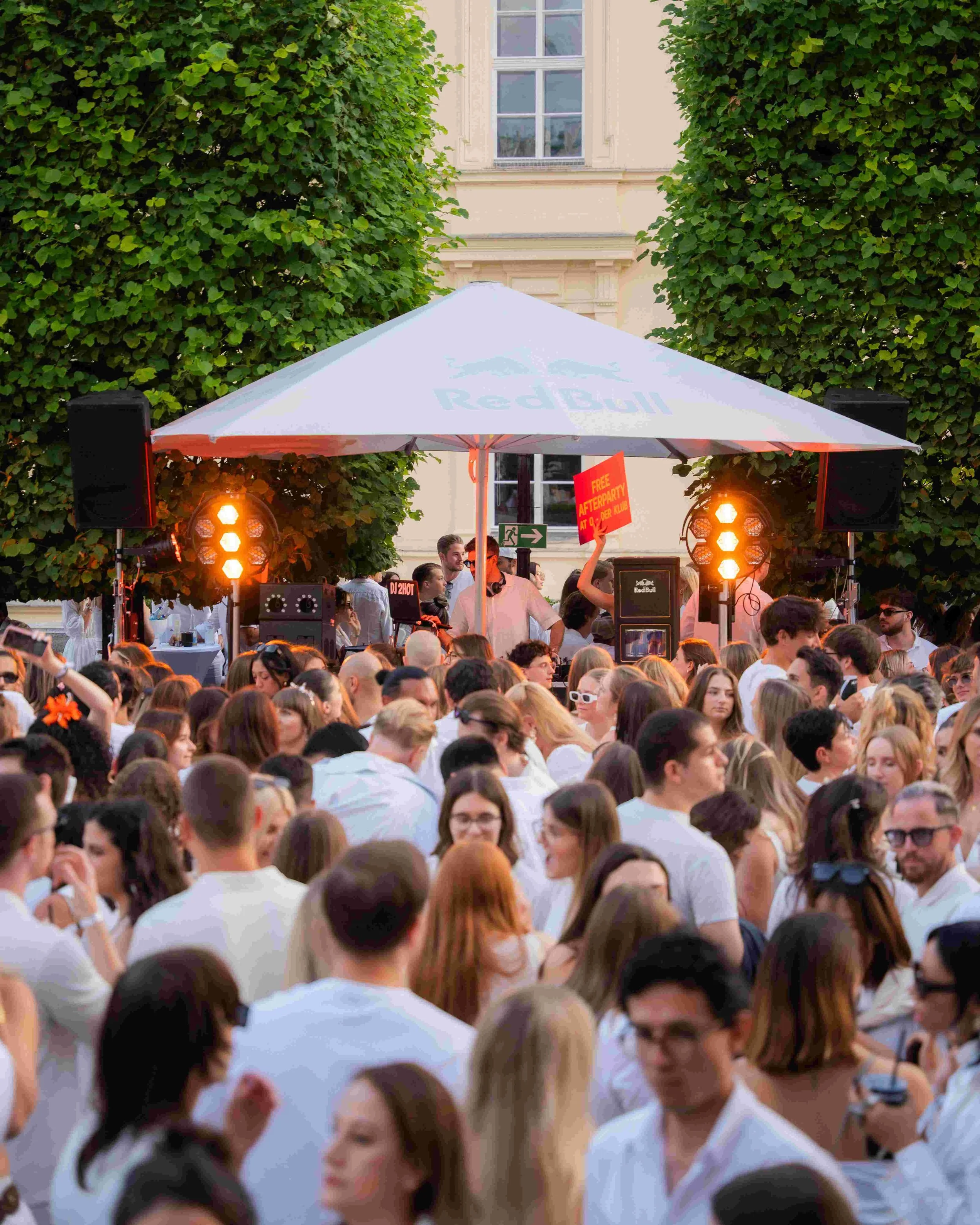 Crowd of people attending an outdoor event under a Red Bull branded tent, with large green trees and a building with windows in the background. There are speakers and lights set up for entertainment.