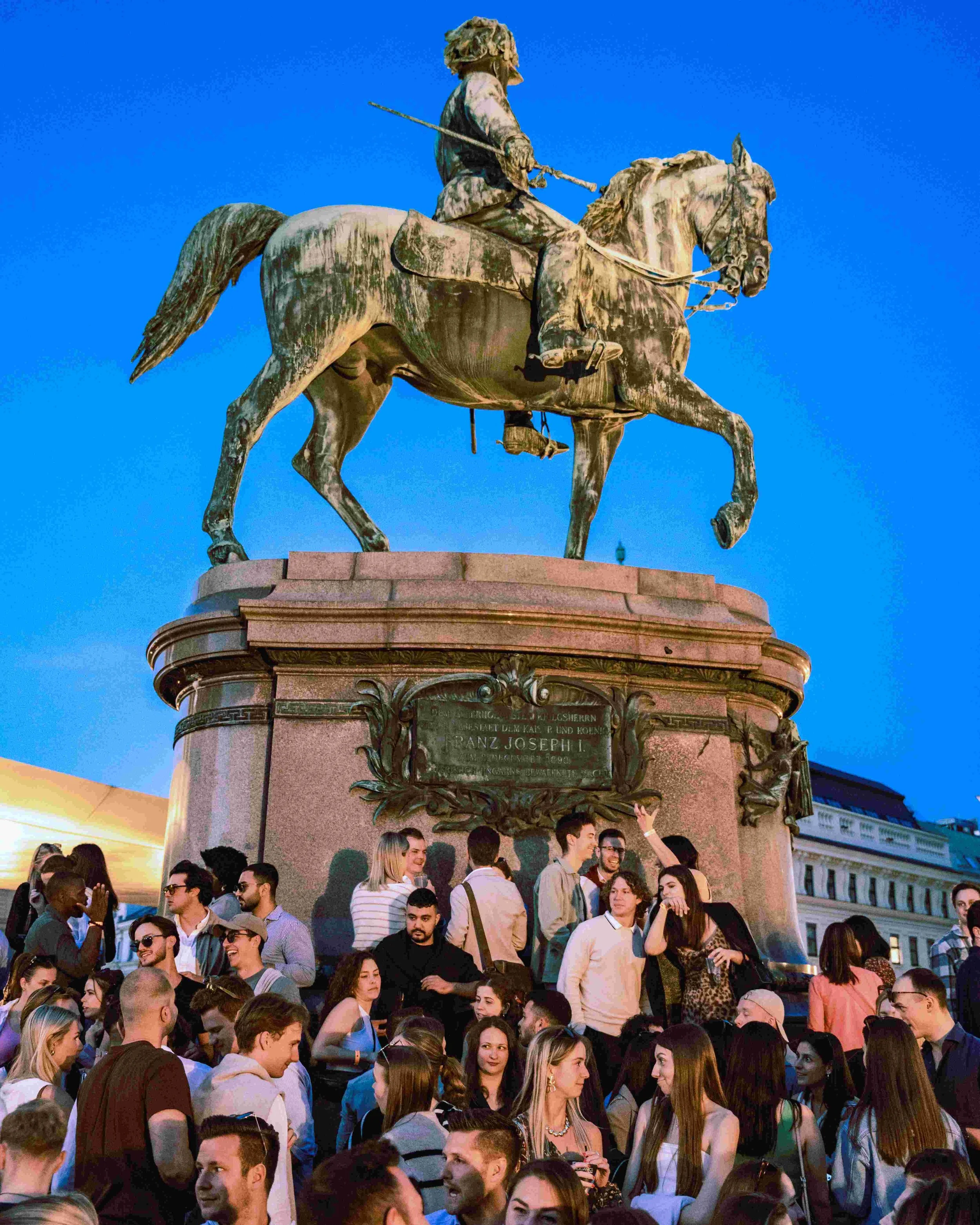 Crowd of people gathered around an equestrian statue of a man on a horse, set against a blue sky in an urban area.