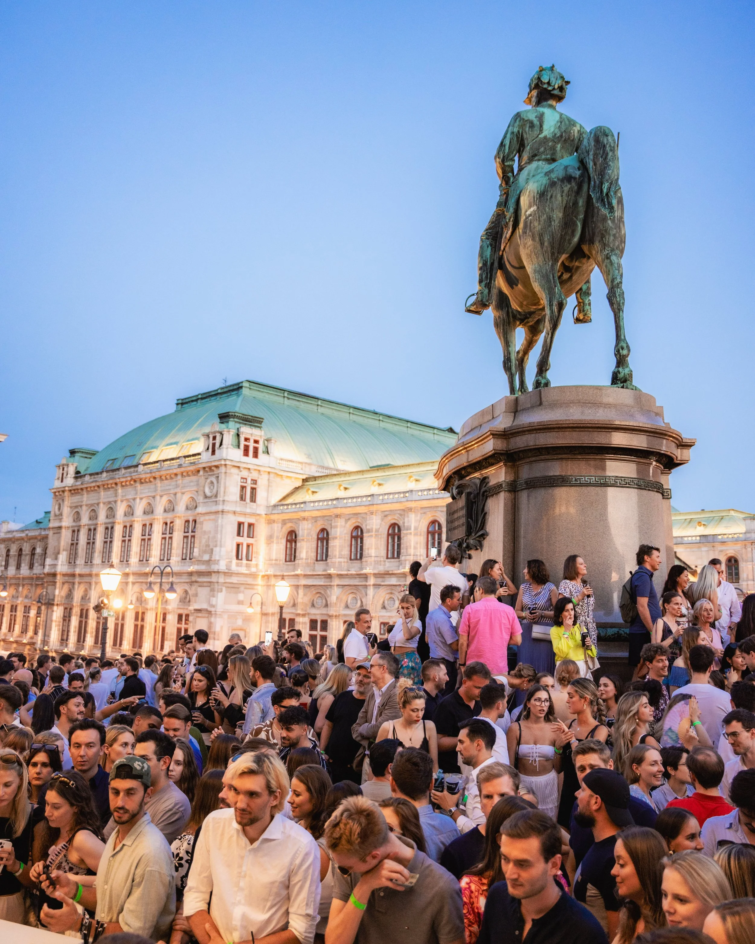 A large crowd of people gathered outdoors near a historic building with a green dome in the background. There is an equestrian statue of a historical figure above them.