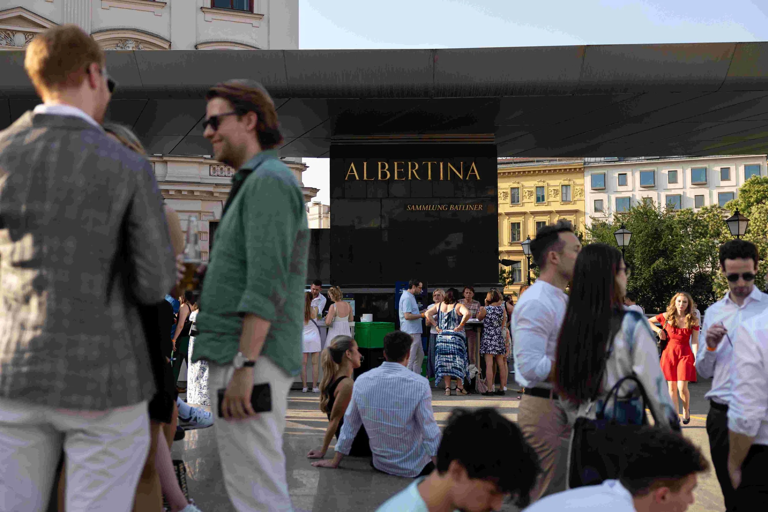 People gathered outdoors on a sunny day in front of a building with European architecture. Some people are standing, and others are sitting or kneeling on the ground. Prominent signage reads 'ALBERTINA' and 'SAMMLUNG BATLINER' on a black board.