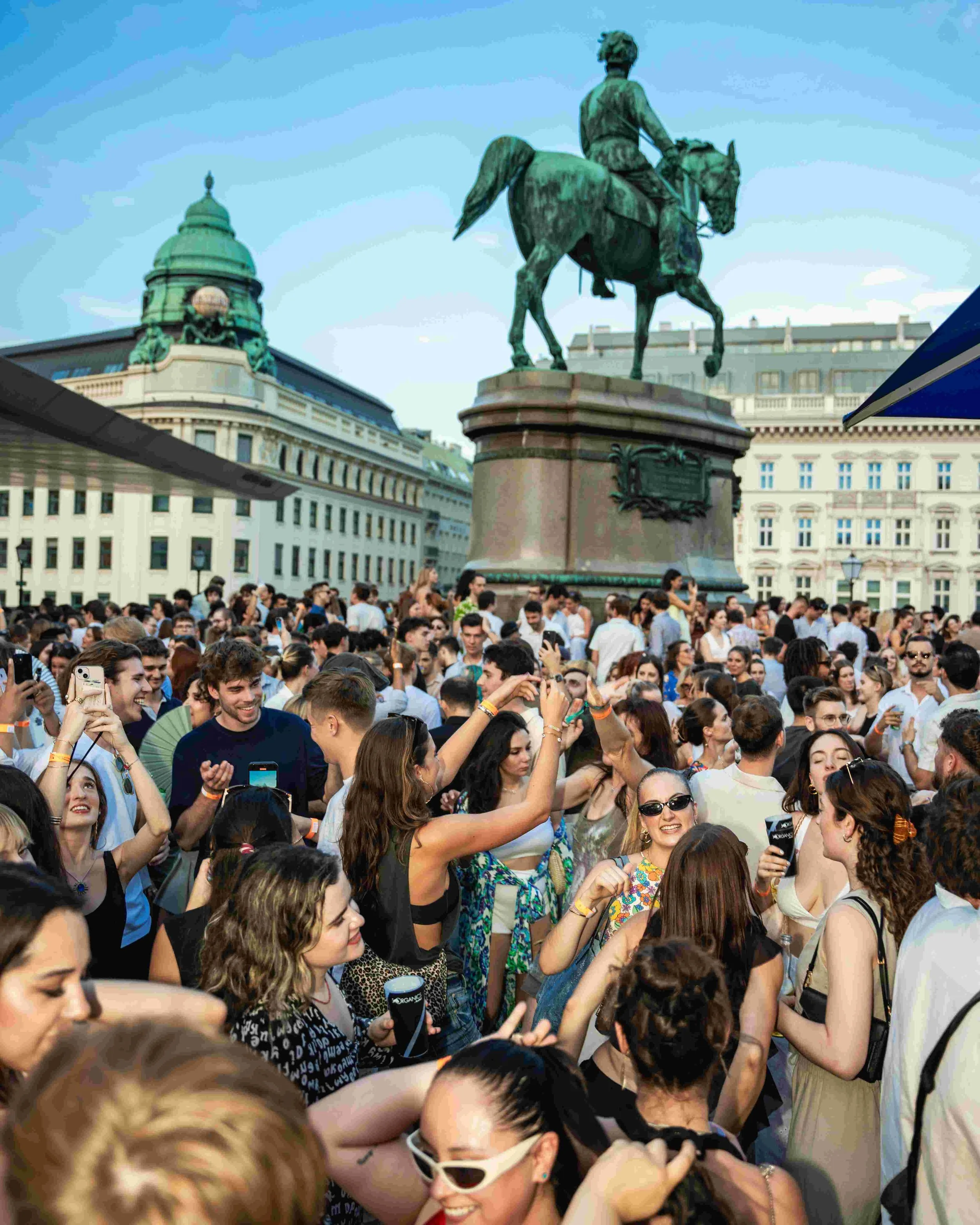 A large crowd of people gathered outdoors, with many taking photos and talking. In the background, there is an equestrian statue of a man in uniform and historical buildings, indicating a city square or public gathering area.