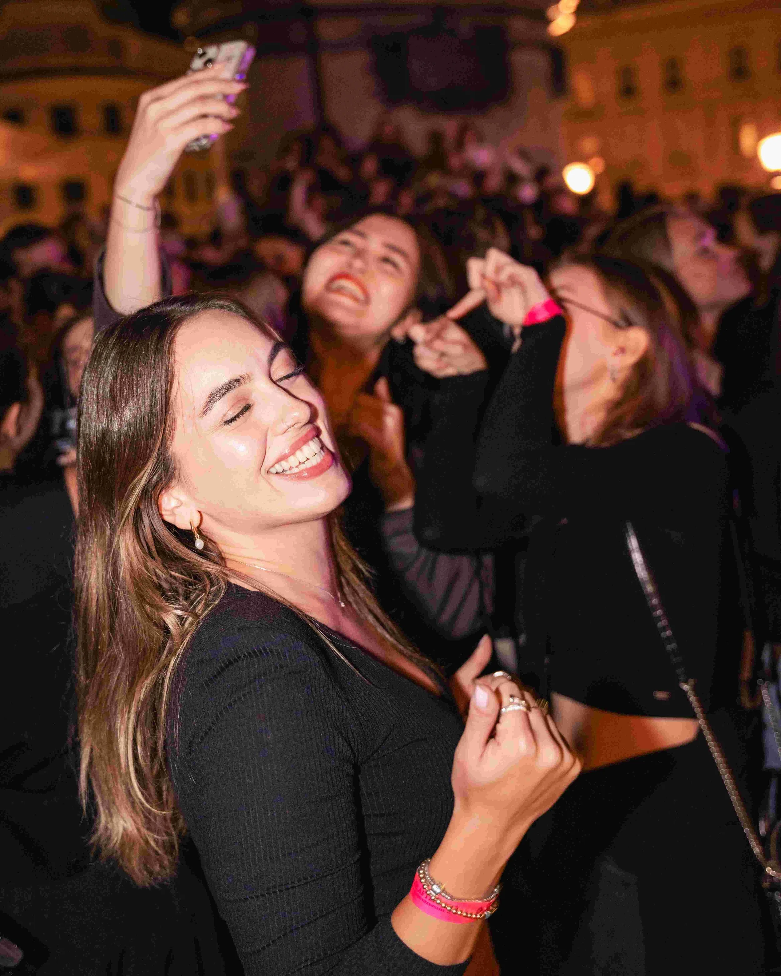 A woman with long, wavy brown hair smiling with eyes closed, wearing earrings, a black long-sleeved top, and bracelets, taking a photo or recording with her smartphone in a crowded indoor setting with warm lighting and blurred people in the backgroun