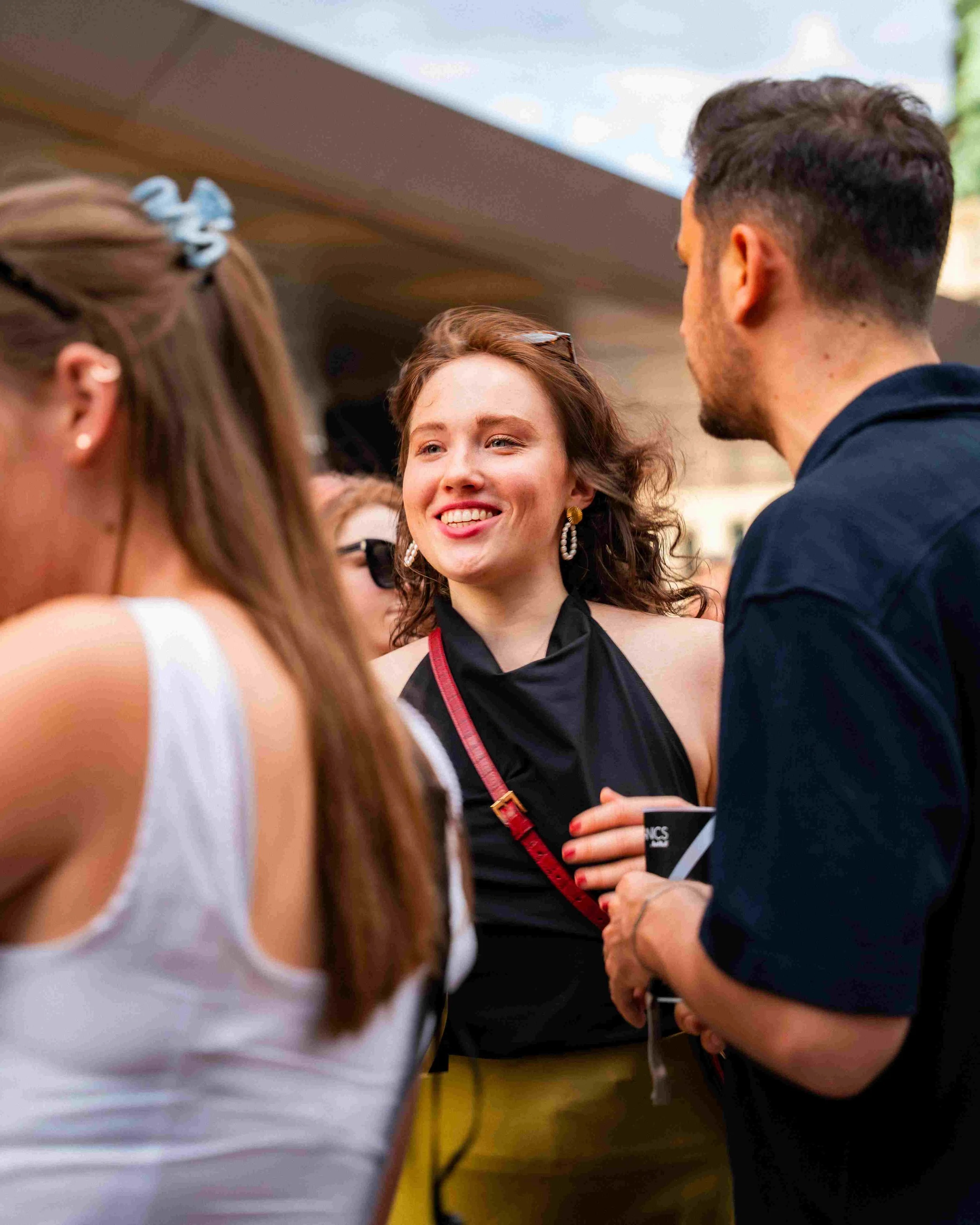 A group of people chatting at a social event, with a woman smiling in the center, wearing earrings and a black top, and a man in a dark shirt facing her. Other people are partially visible in the foreground and background.