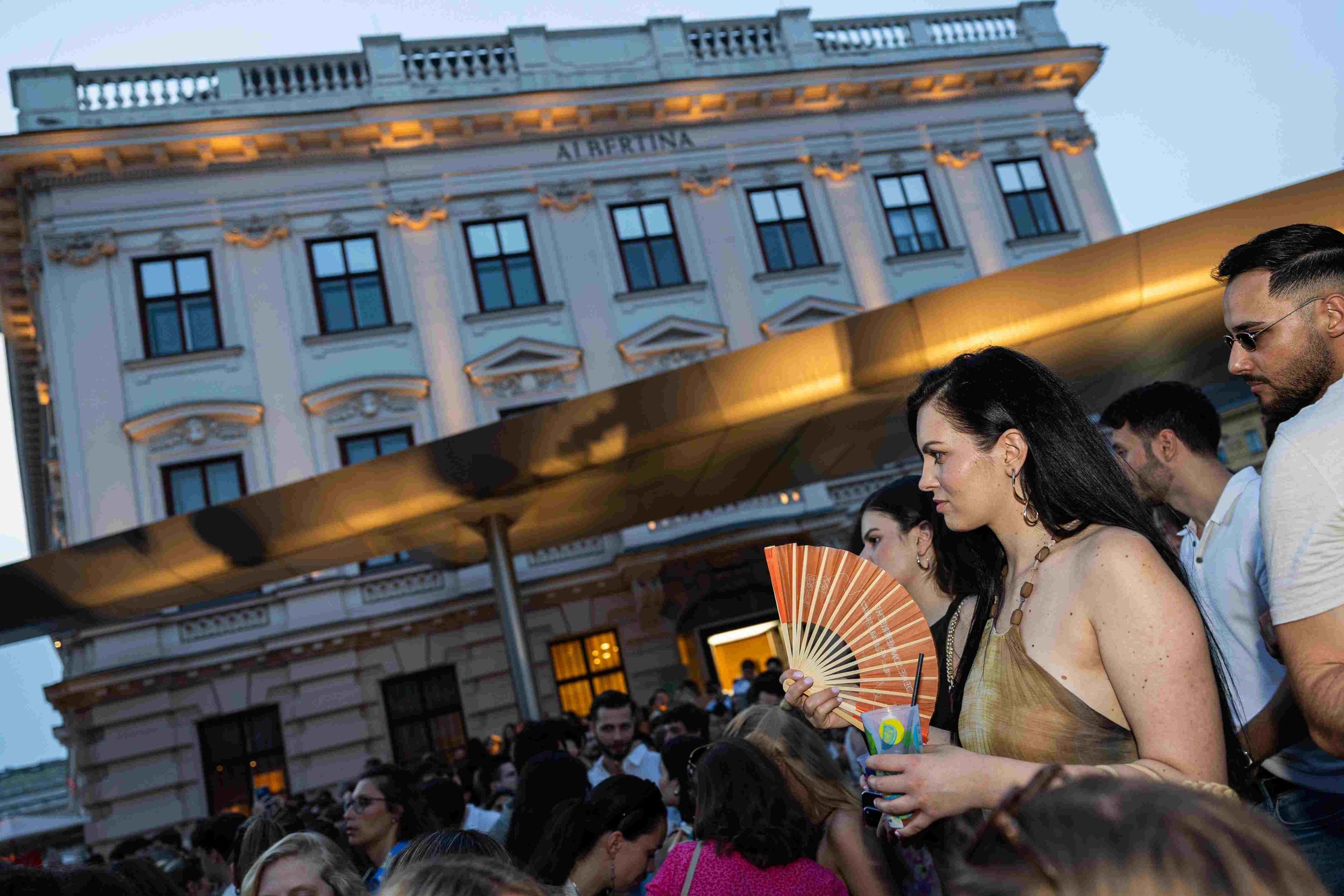 People gathered outdoors in an evening setting with a historical building in the background. A woman in the foreground holds a paper fan and wears a necklace, while others around her are engaged in conversation or looking around.